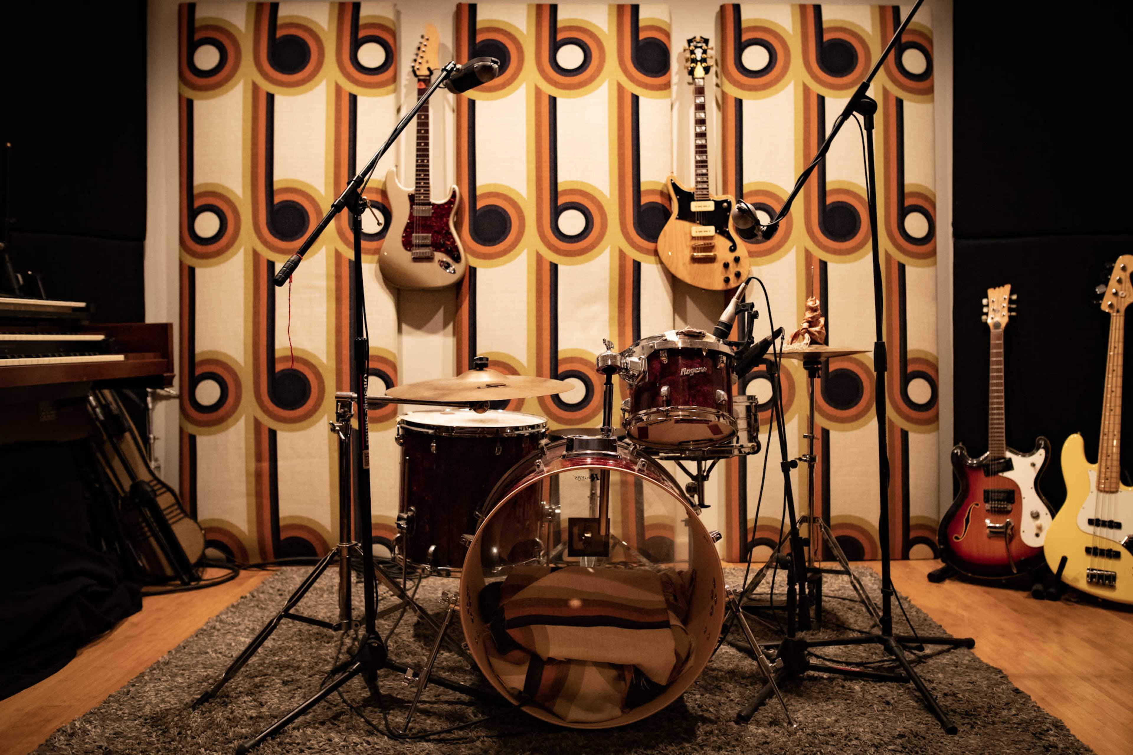 A drum set is positioned in a music studio surrounded by various guitars and a patterned backdrop.