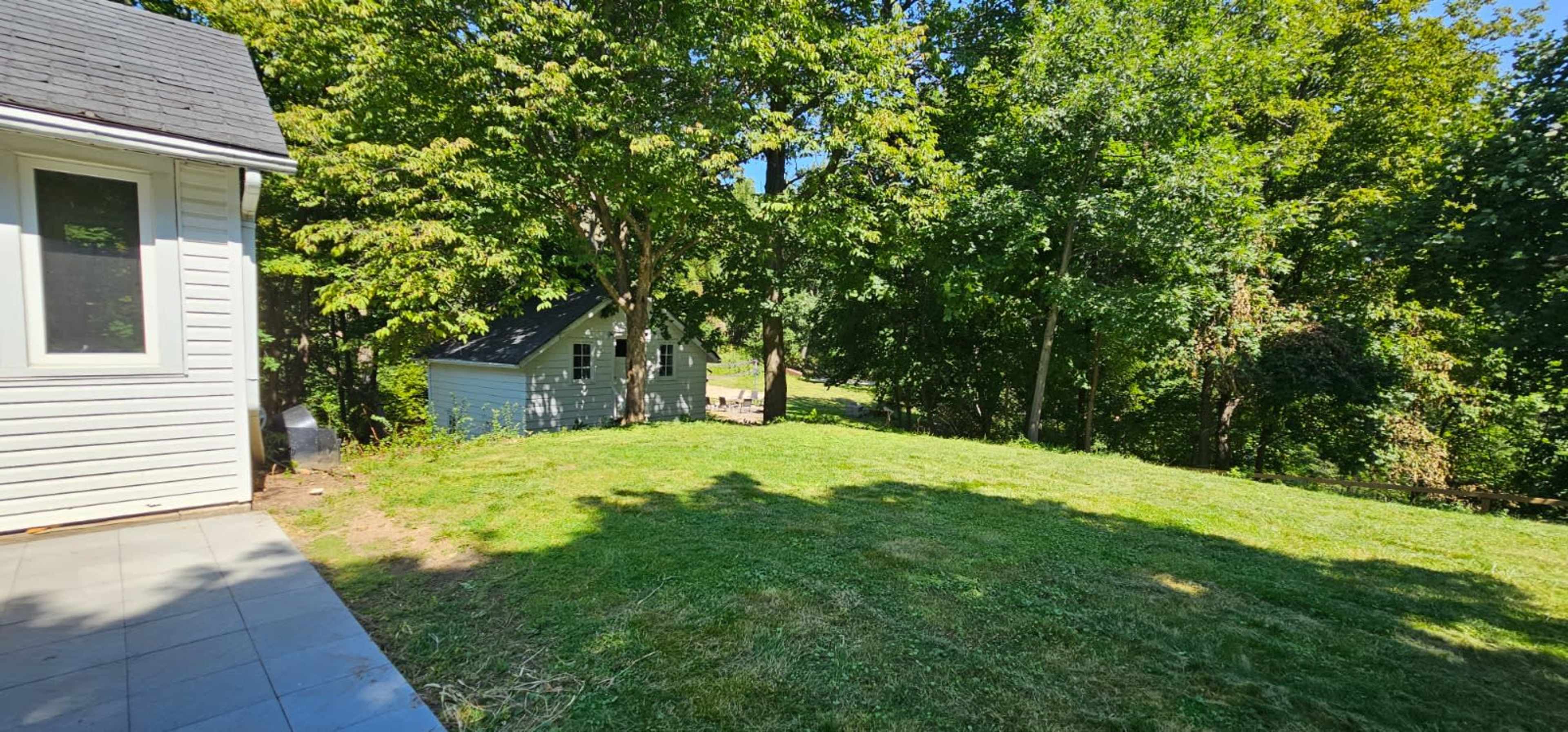 The image shows a grassy yard with trees and a small building in the background, taken from the side of a house.
