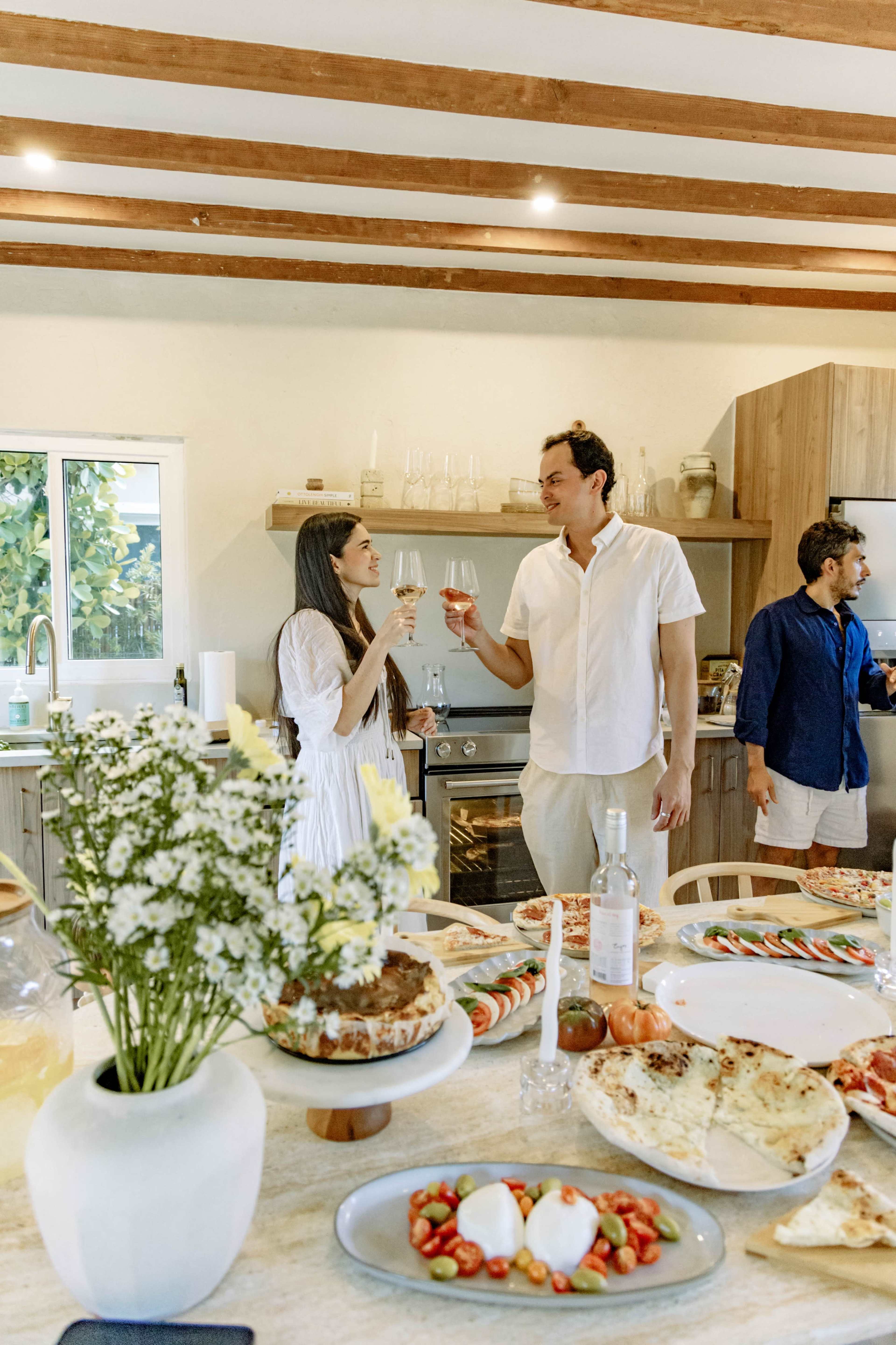 A couple toasts with glasses of wine in a modern kitchen, while another person prepares food in the background.