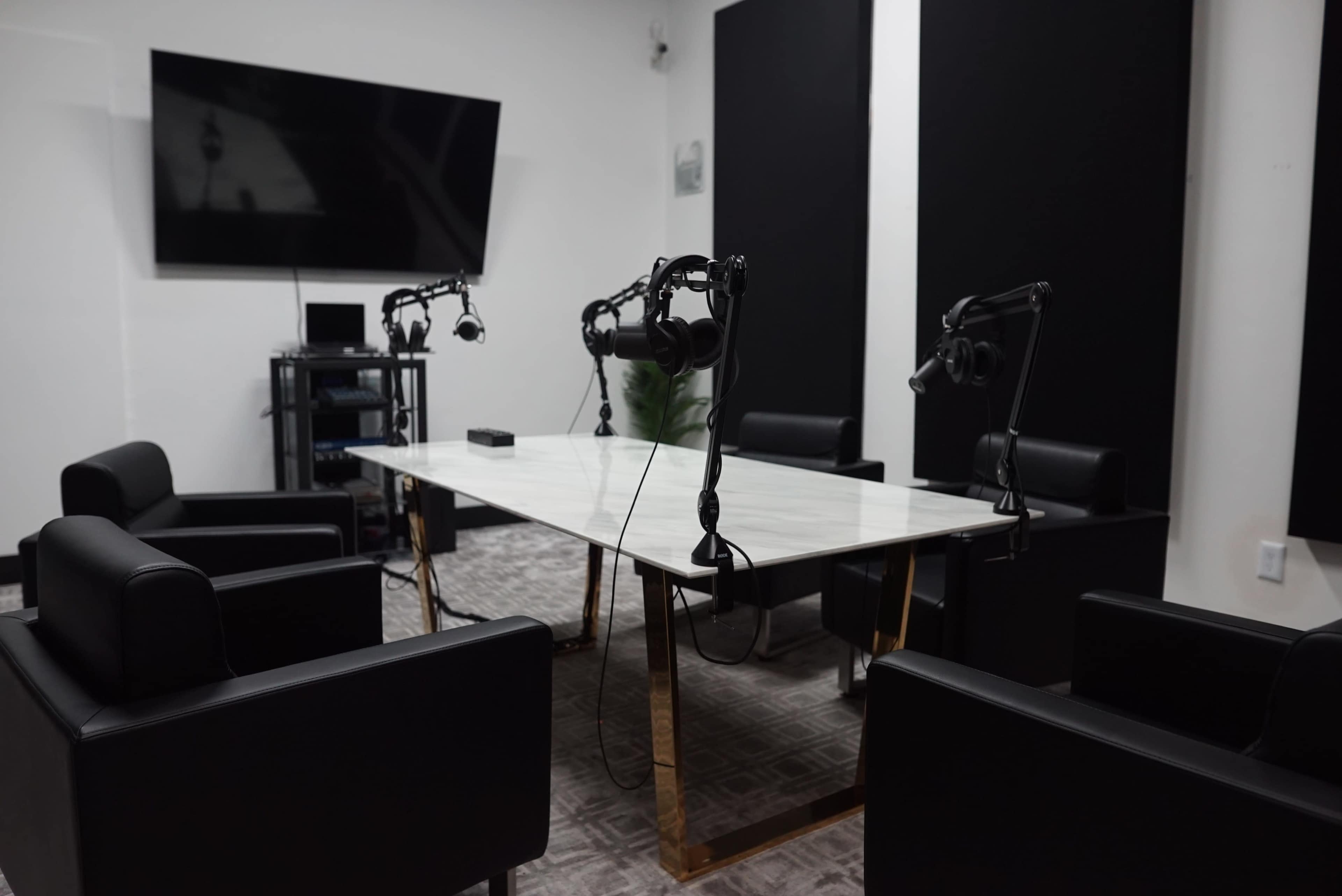 A modern podcast studio features a large white table surrounded by black chairs, with several microphone setups and a television on the wall.