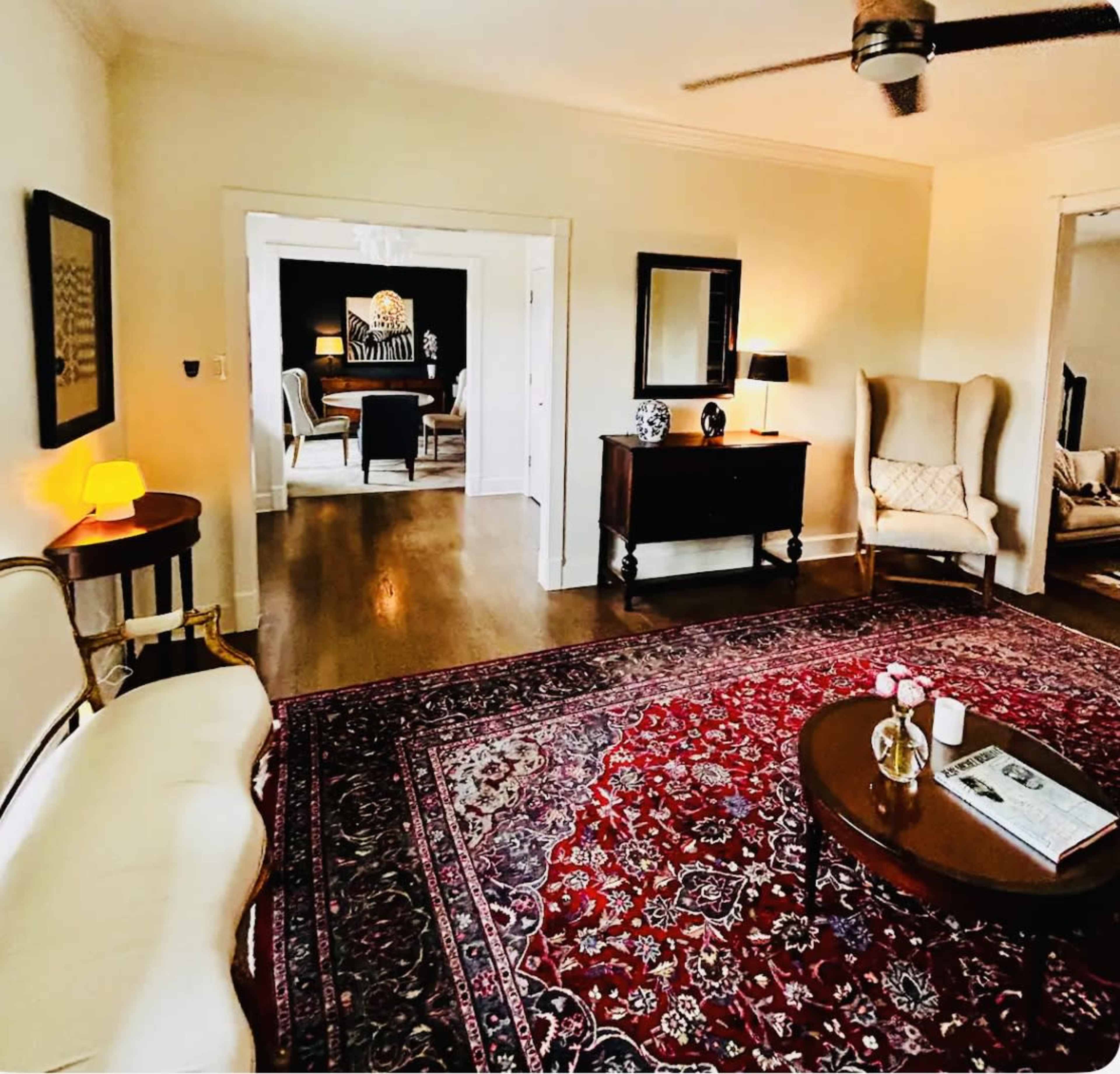 A cozy living room featuring a patterned rug, a wooden coffee table with decorative items, and a sofa, leading to an adjacent room with additional seating.