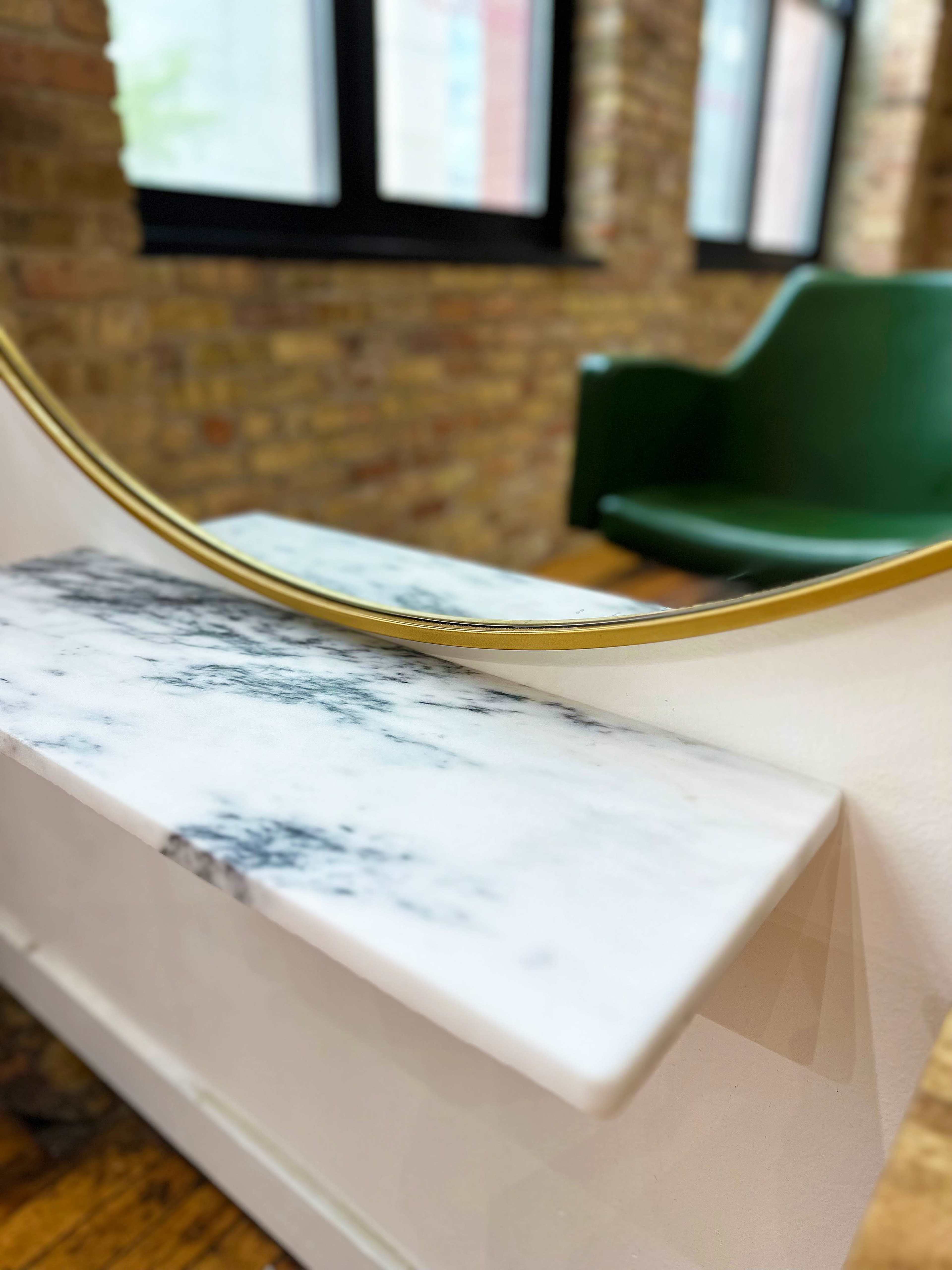 A round mirror with a gold frame reflects a marble shelf positioned in front of a green chair and a brick wall.
