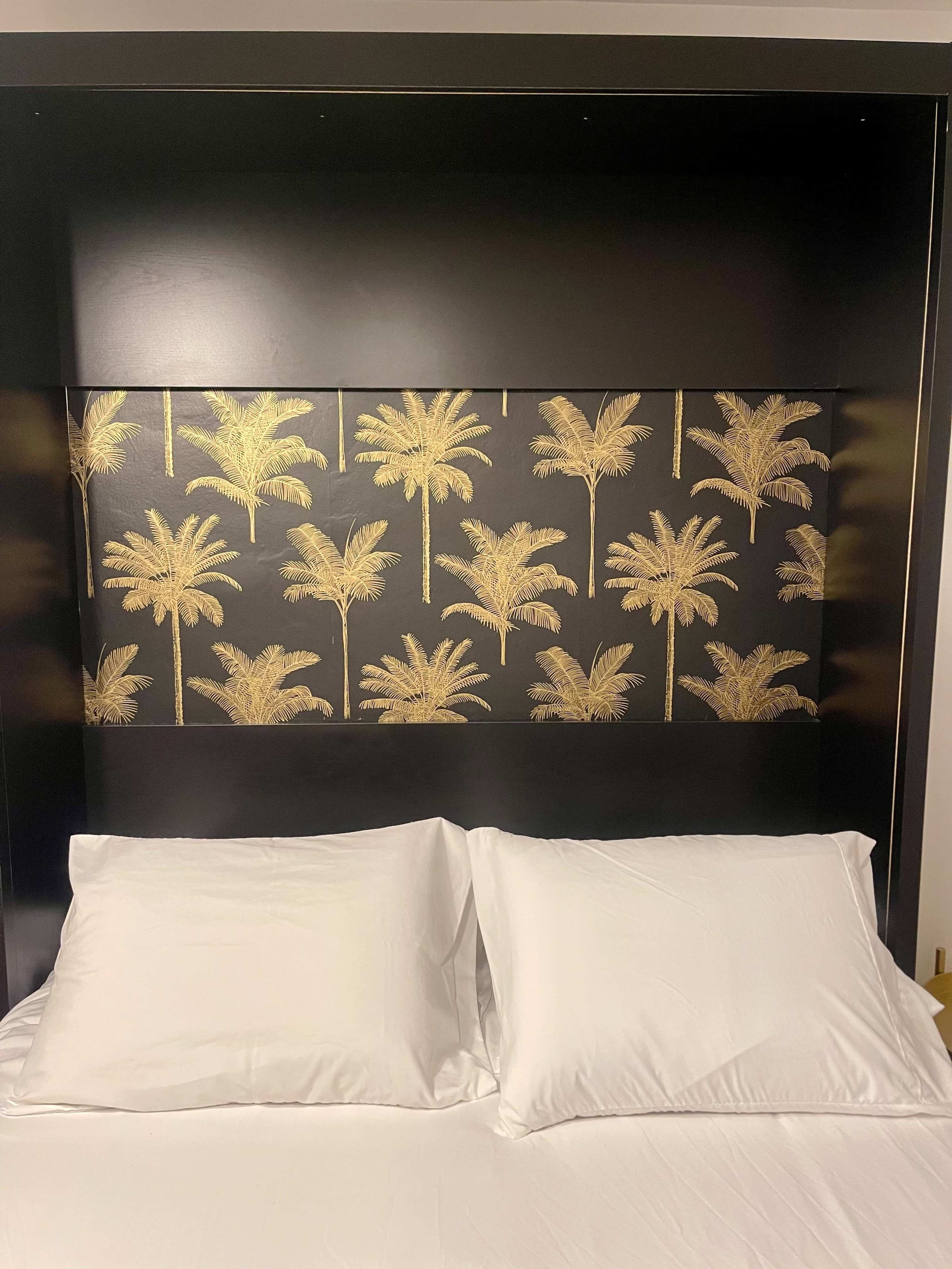 The image shows a bed with two white pillows against a black headboard featuring a gold palm tree pattern.