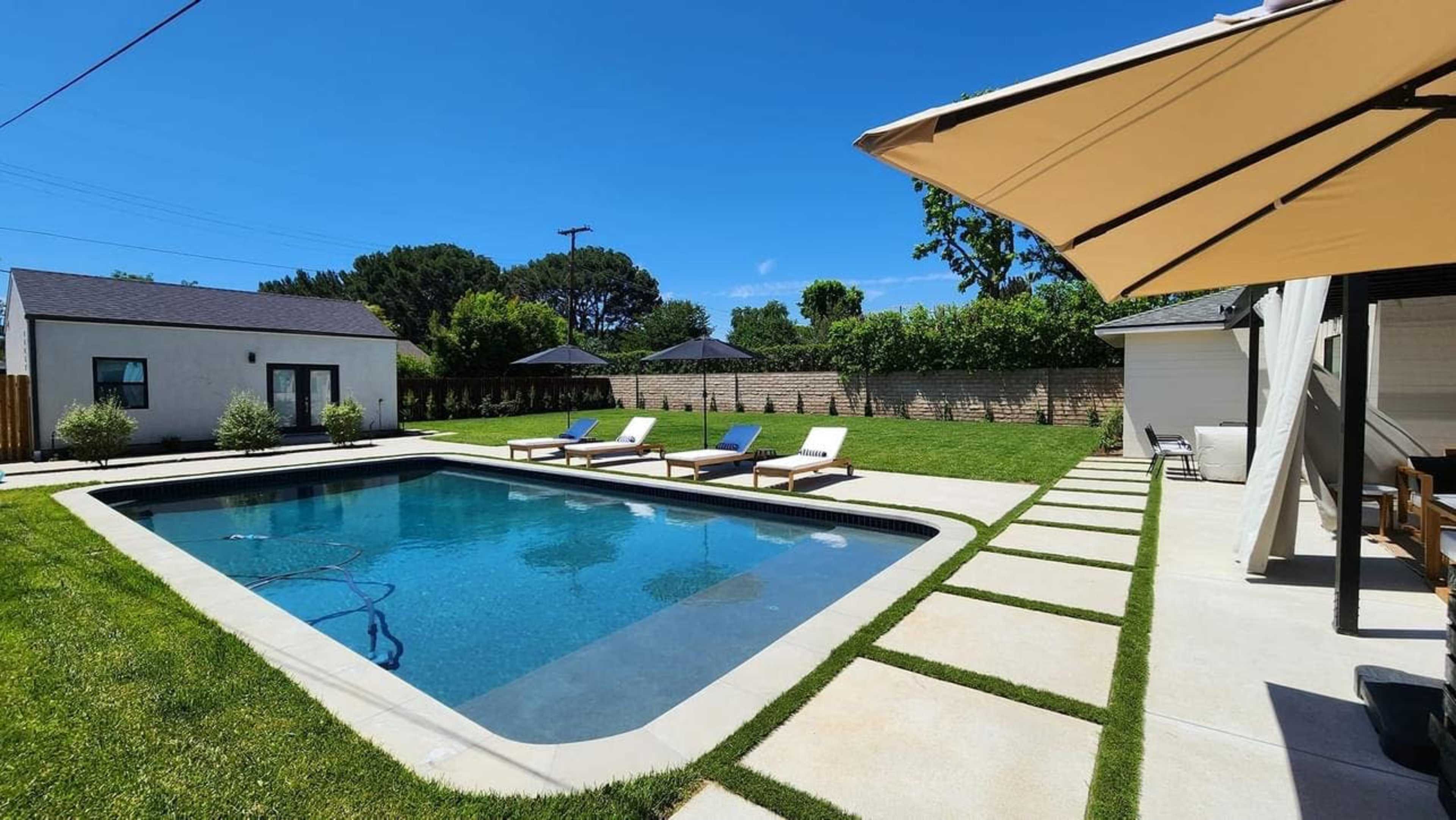 The image shows a rectangular swimming pool surrounded by lounge chairs, a grassy area, and tree-lined landscaping under a clear blue sky.