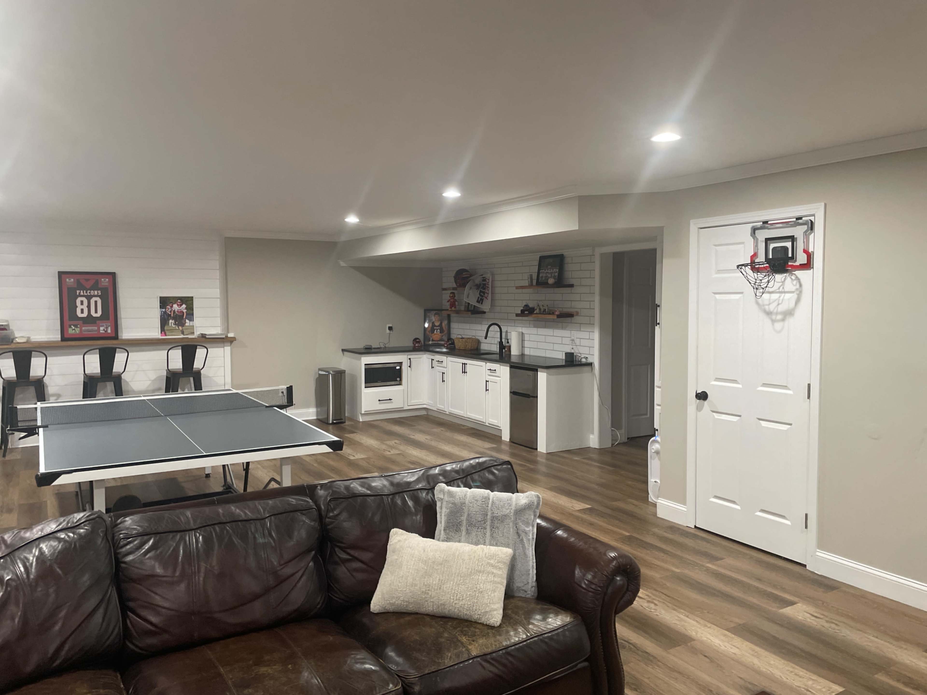 The image shows a finished basement featuring a ping pong table, a kitchenette area, and a seating area with a brown couch.