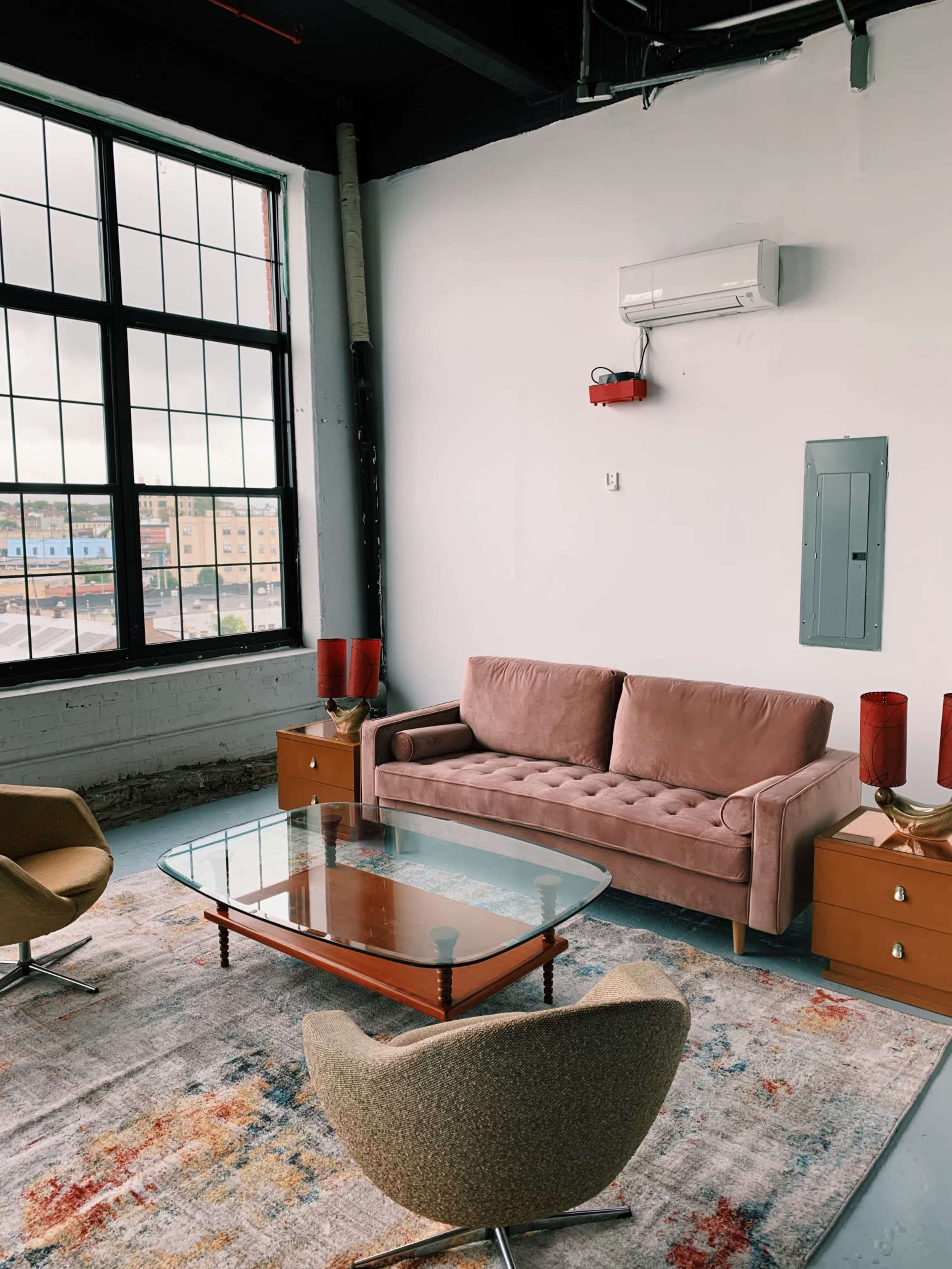 A modern lounge area features a pink sofa, two armchairs, a glass coffee table, and two red lamps against a backdrop of large windows.