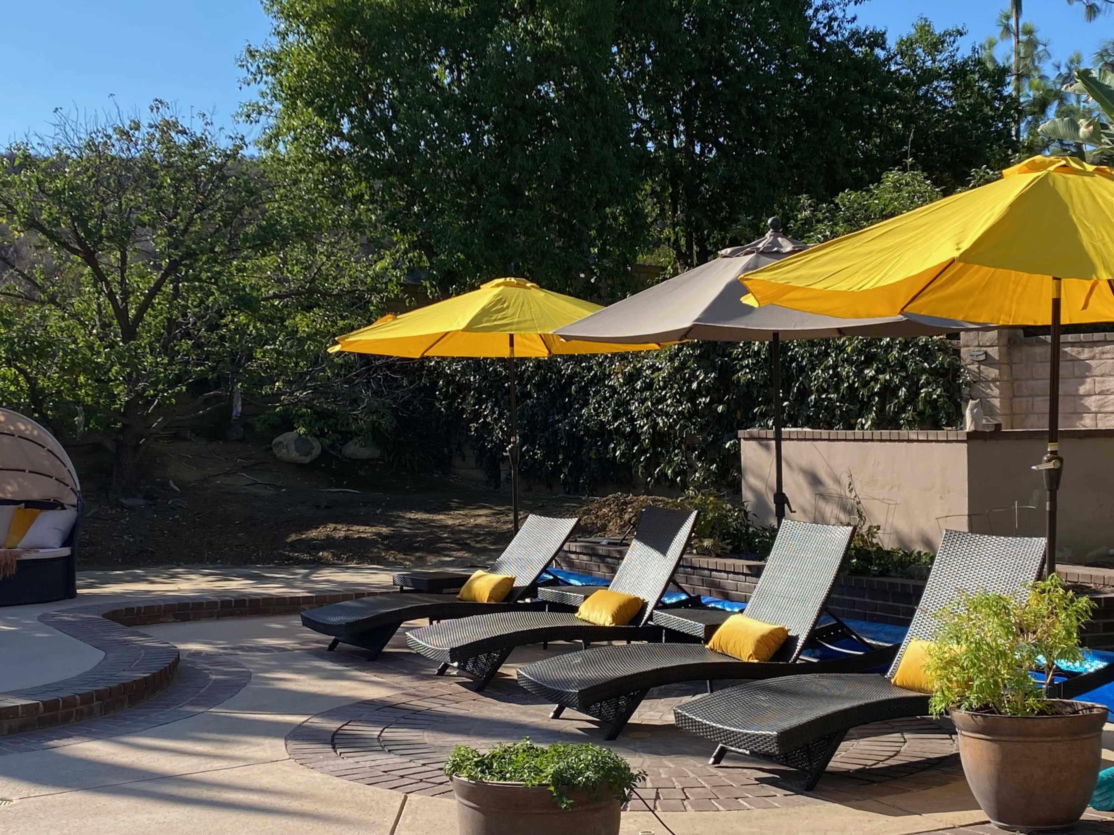 The image shows a poolside area with several lounge chairs, yellow umbrellas, and a small round table surrounded by greenery.