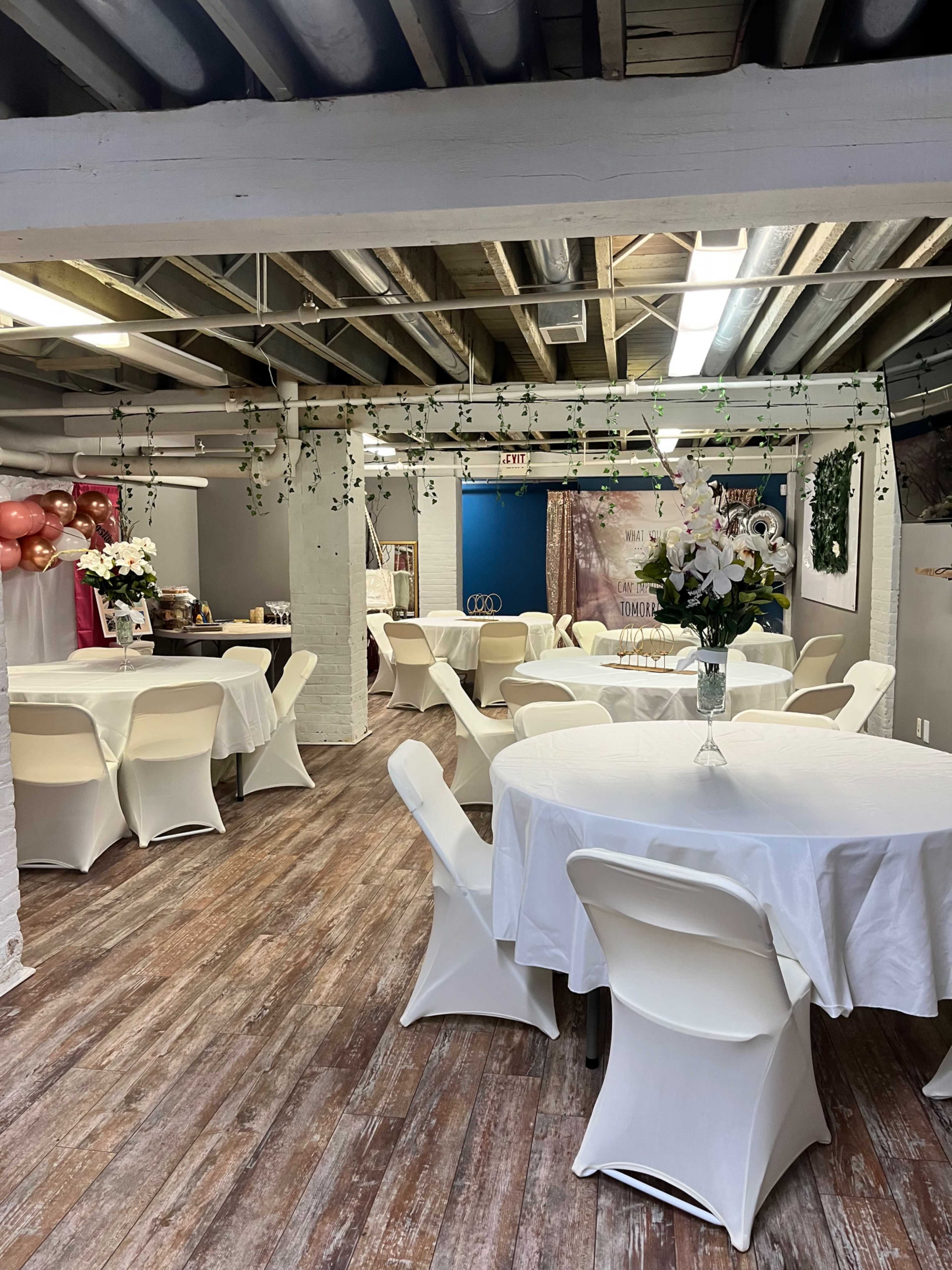 The image shows a spacious event room with several round tables covered in white tablecloths, decorated with vases of flowers, and walls adorned with balloons and streamers.