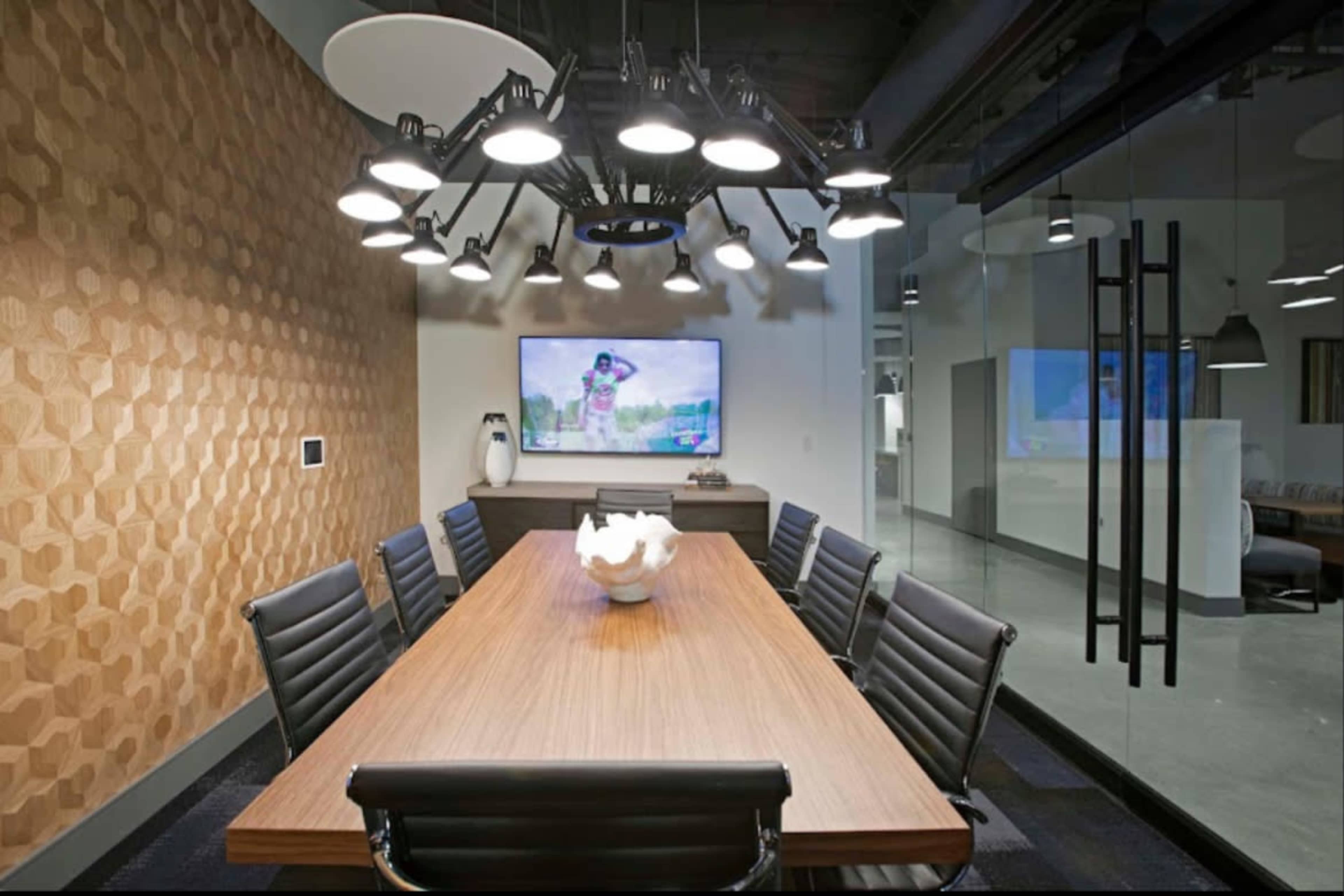 A conference room features a long wooden table surrounded by black chairs, a large screen displaying a presentation, and a modern overhead light fixture.