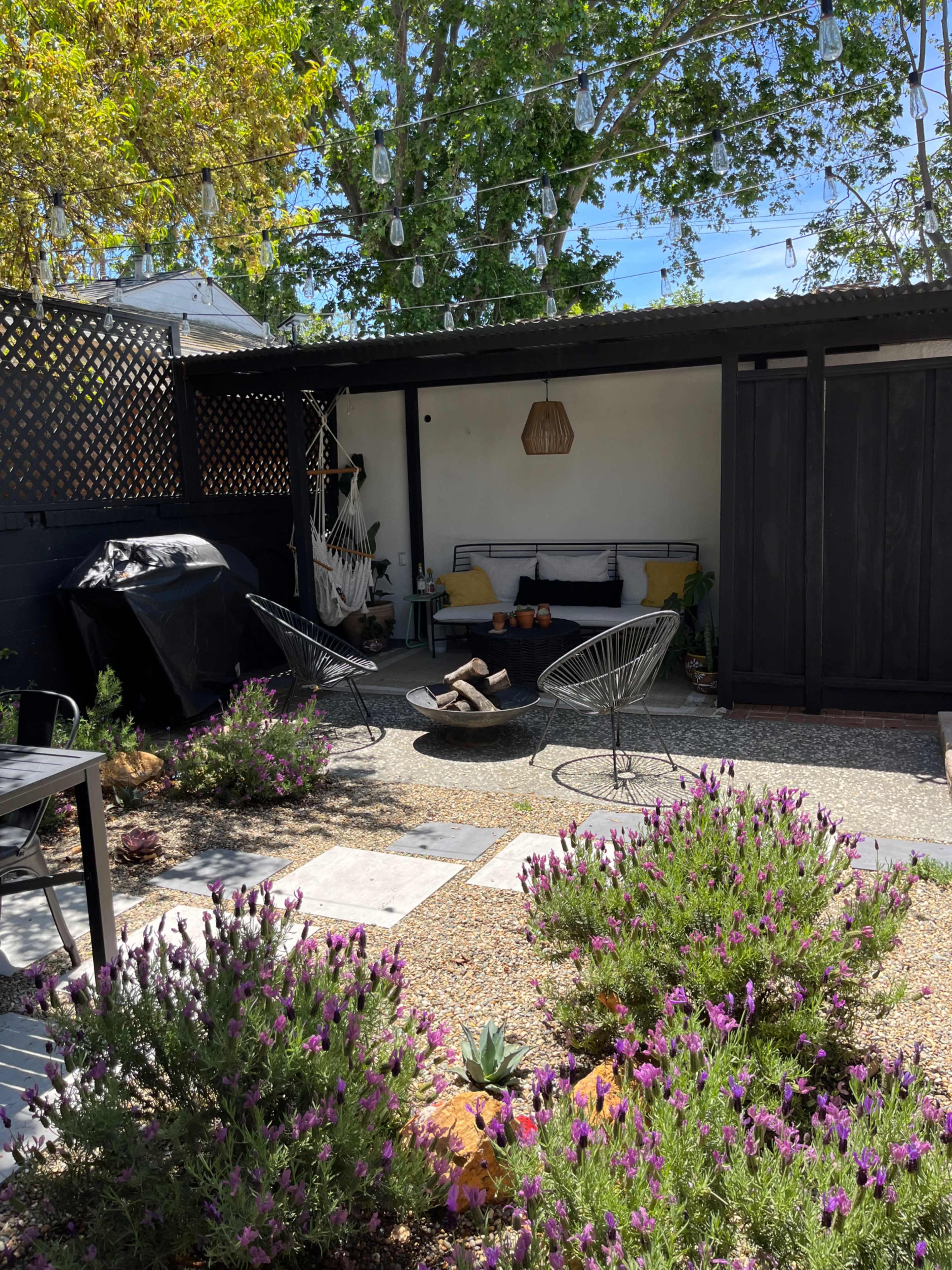 A cozy outdoor seating area features two chairs around a fire pit, surrounded by lavender plants and string lights overhead.