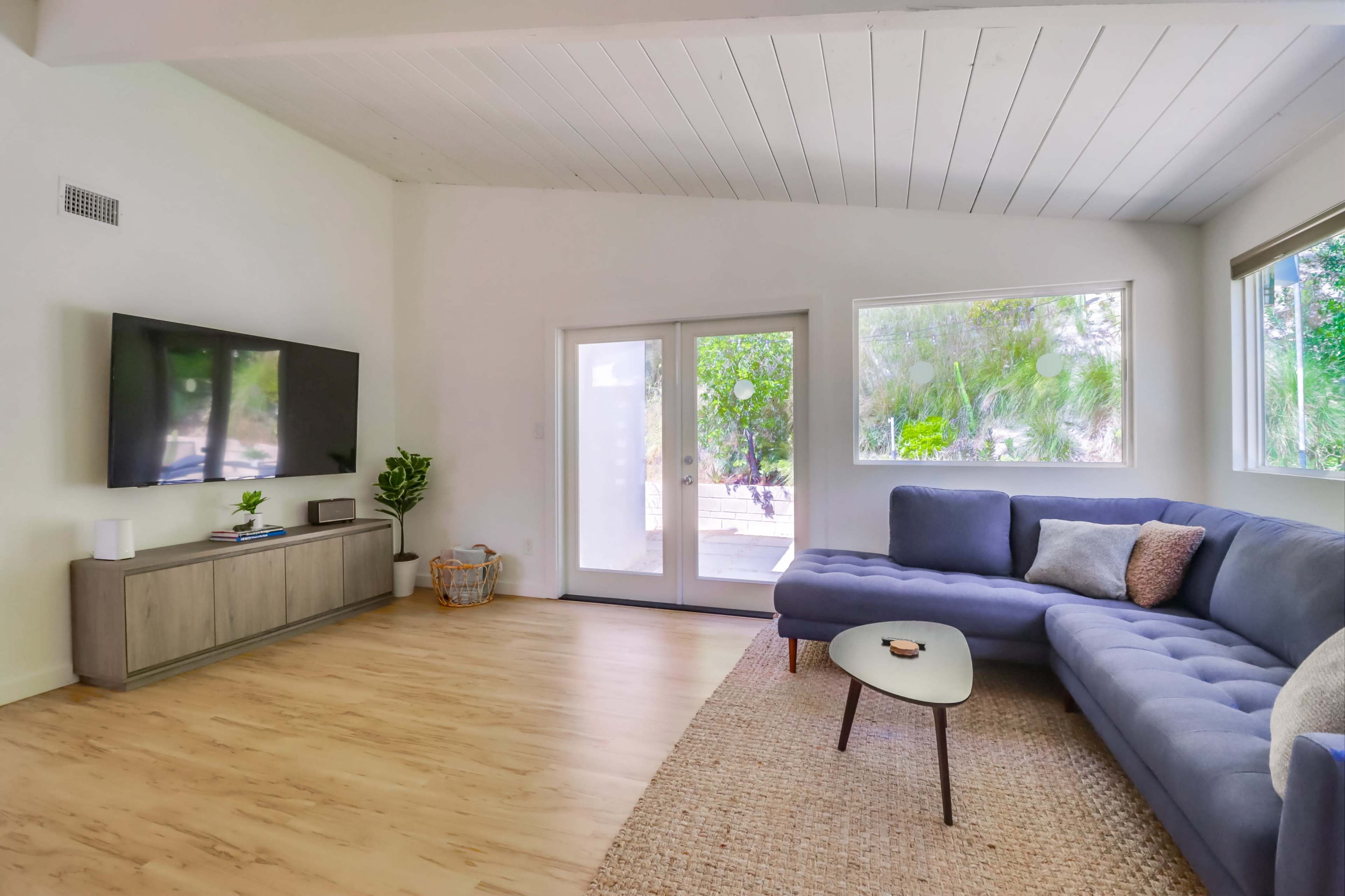 A spacious living room features a blue sectional sofa, a modern coffee table, a television mounted on the wall, and large windows that overlook greenery outside.