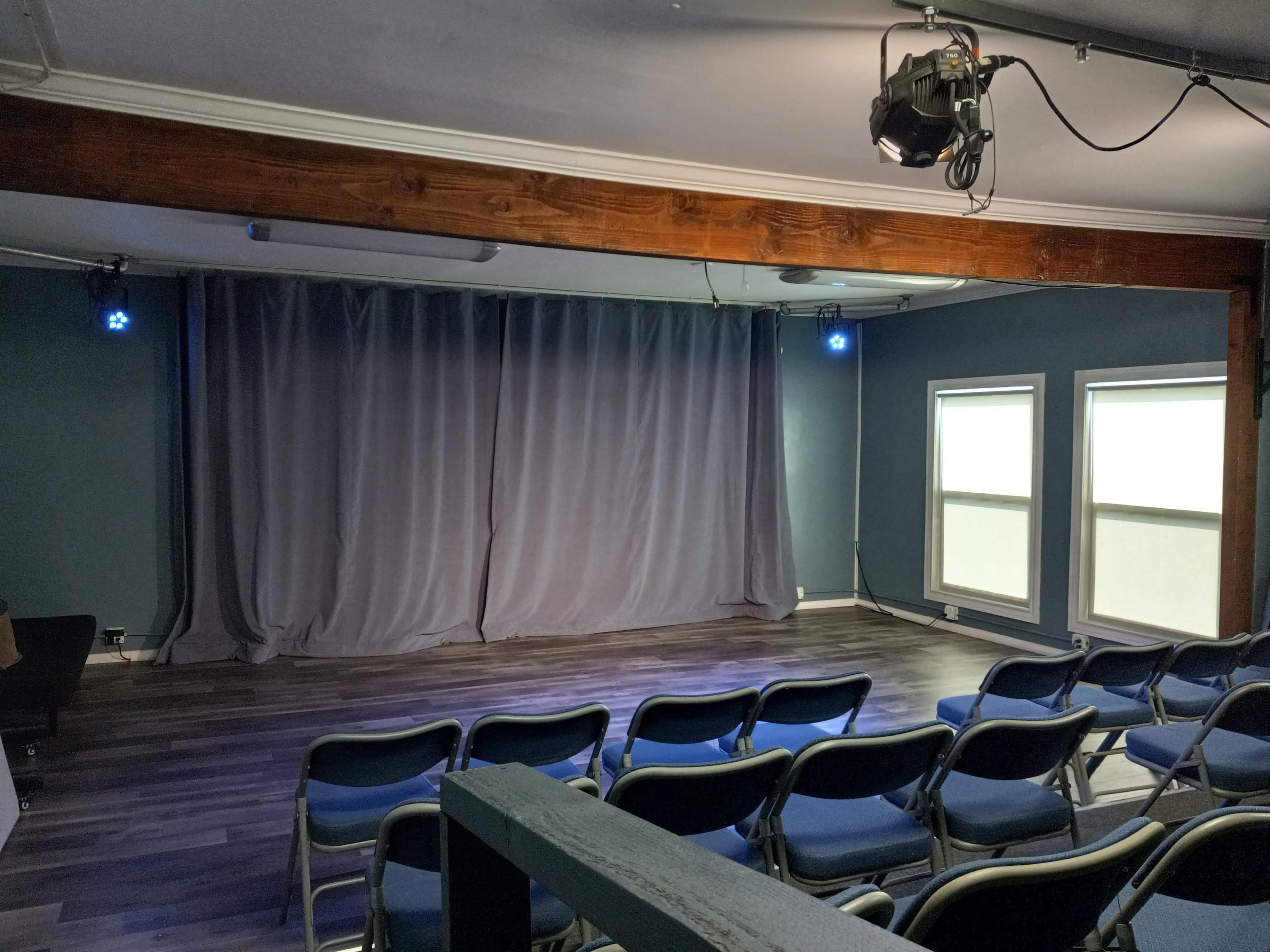 A small performance space with a gray curtain, blue stage lights, and rows of blue chairs facing the stage.