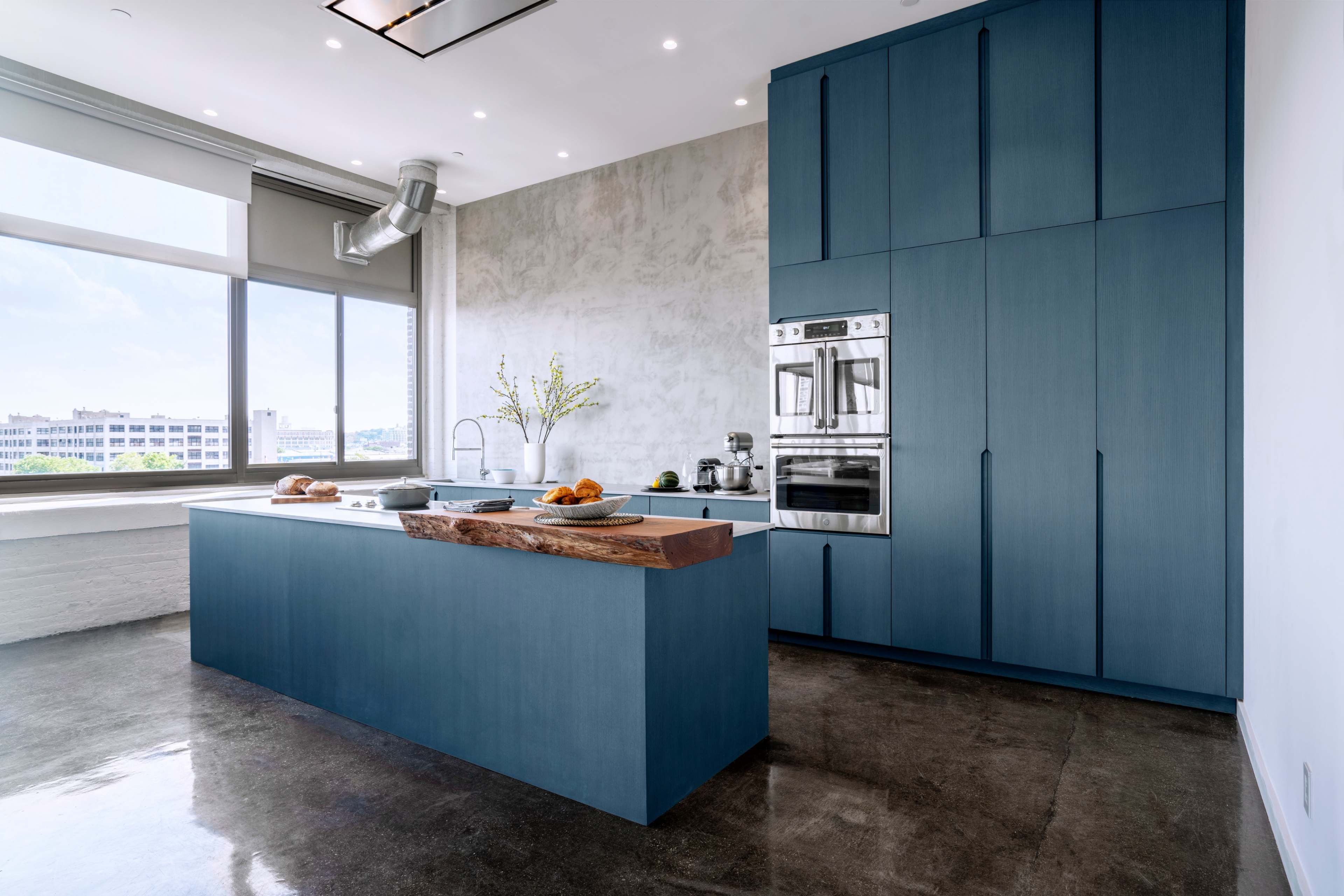 A modern kitchen features a large blue island with a wooden countertop, stainless steel appliances, and a concrete wall.