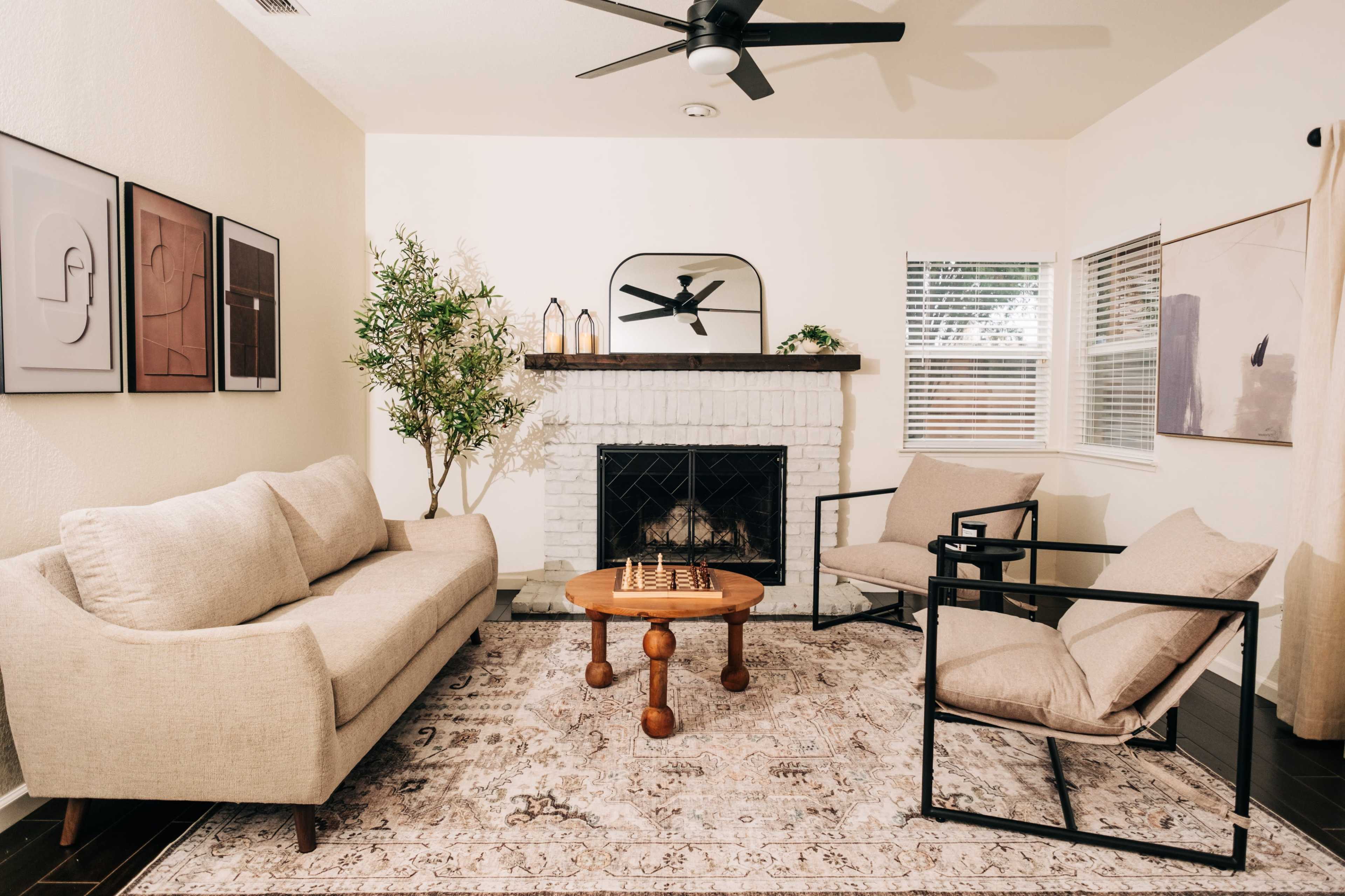 A living room features a beige sofa, two black framed chairs, a wooden coffee table with a chess set, and a fireplace surrounded by white brick.