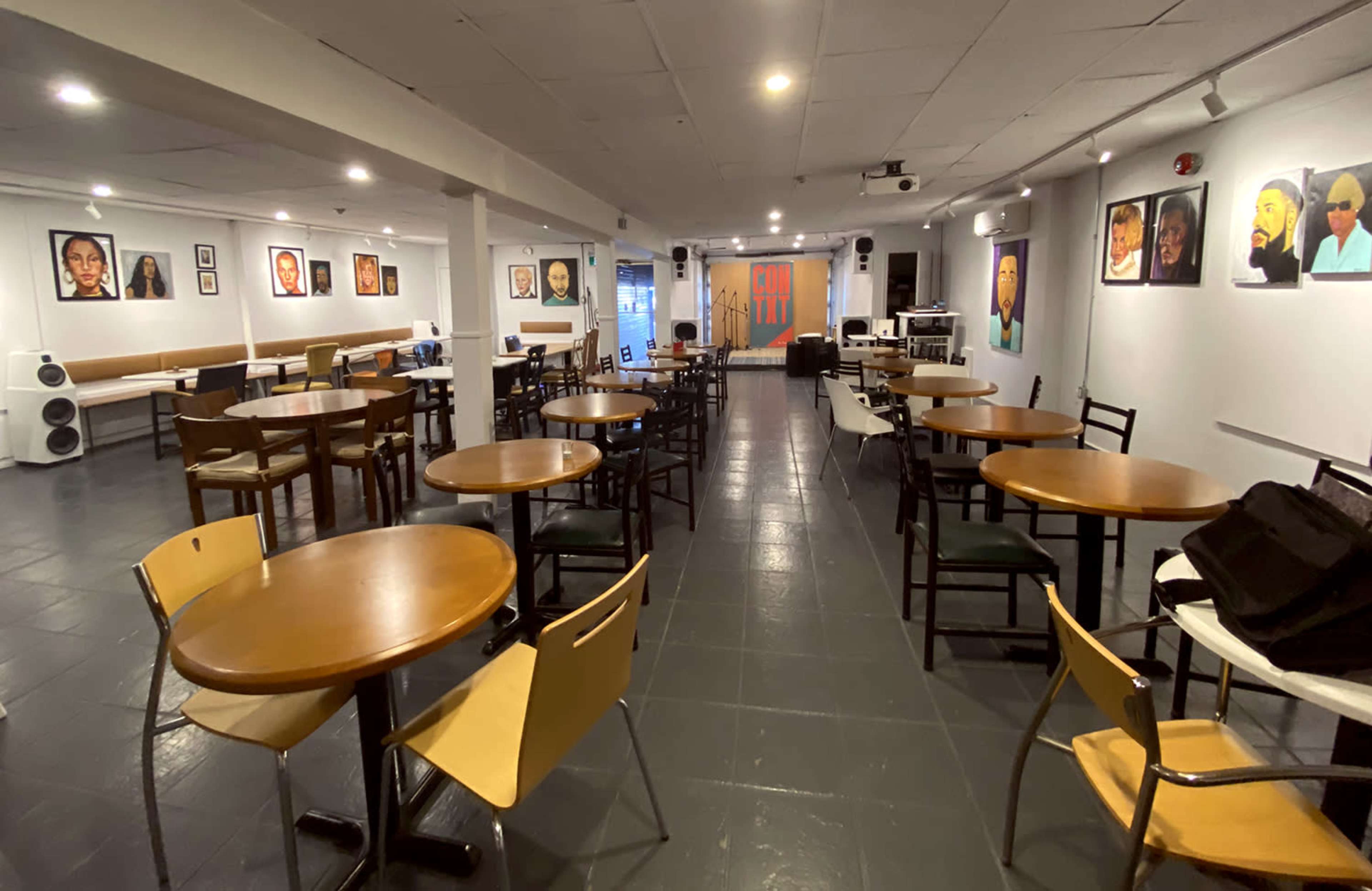 The image shows a spacious indoor café or event space with tables and chairs arranged around the room, featuring artwork on the walls.