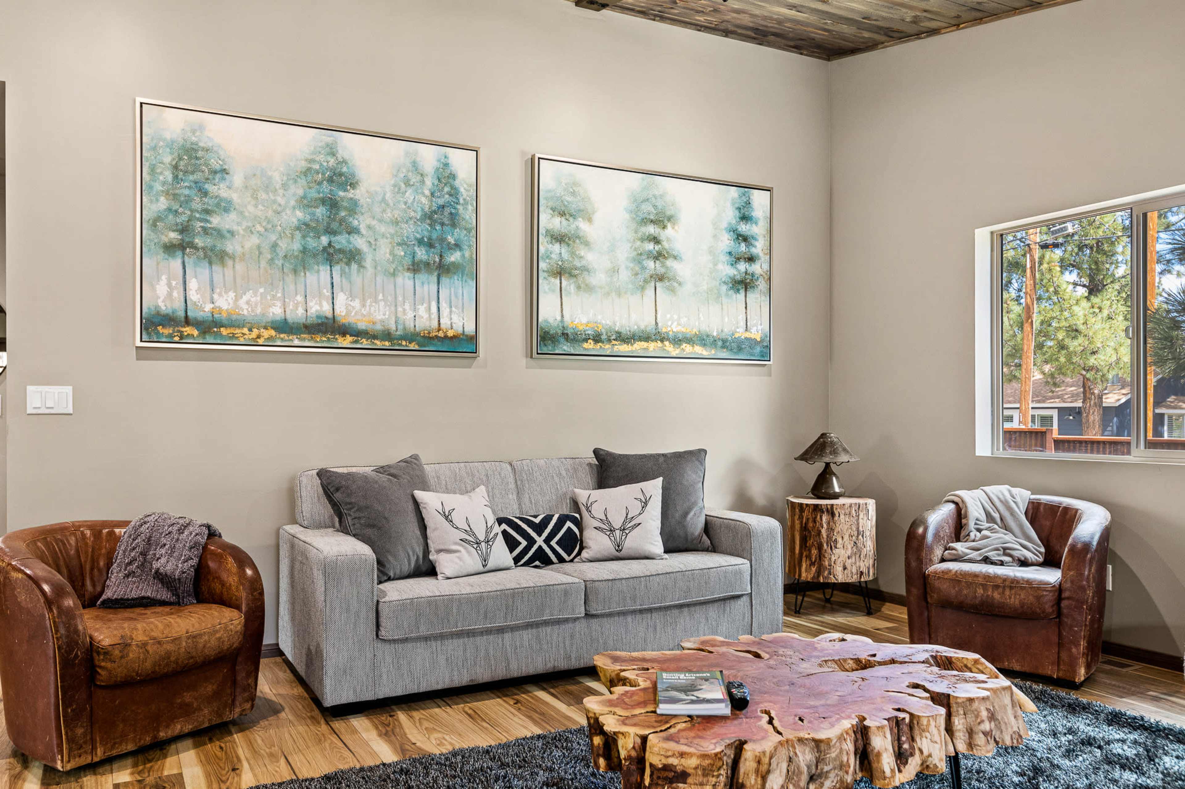 The image features a living room with a gray sofa, two brown chairs, a wooden coffee table, and framed landscape paintings on the wall.