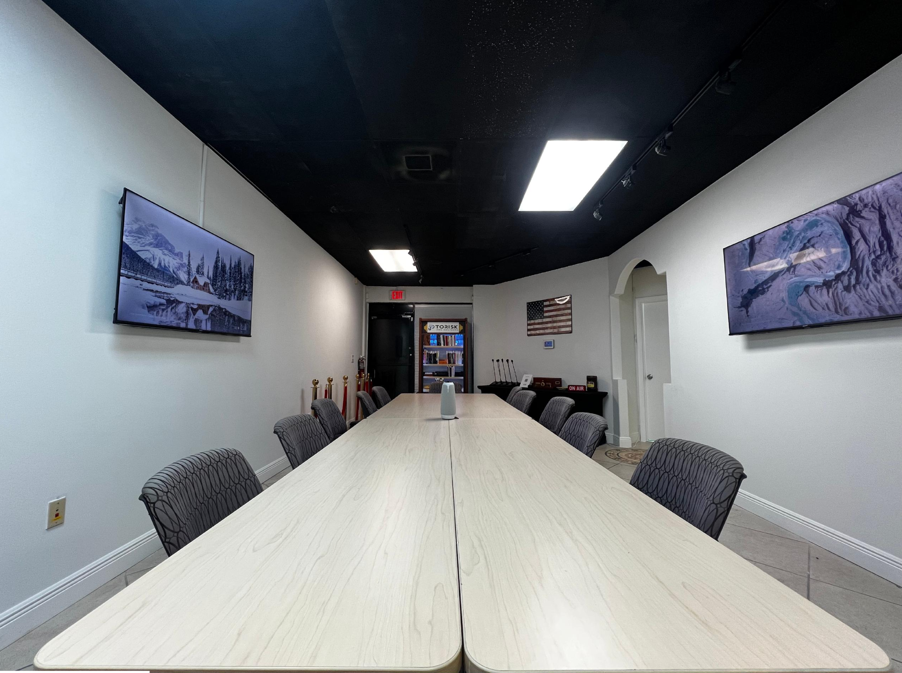 A long, minimalist conference table is centered in a brightly lit room, flanked by two large wall-mounted screens and decorated with framed landscape art.