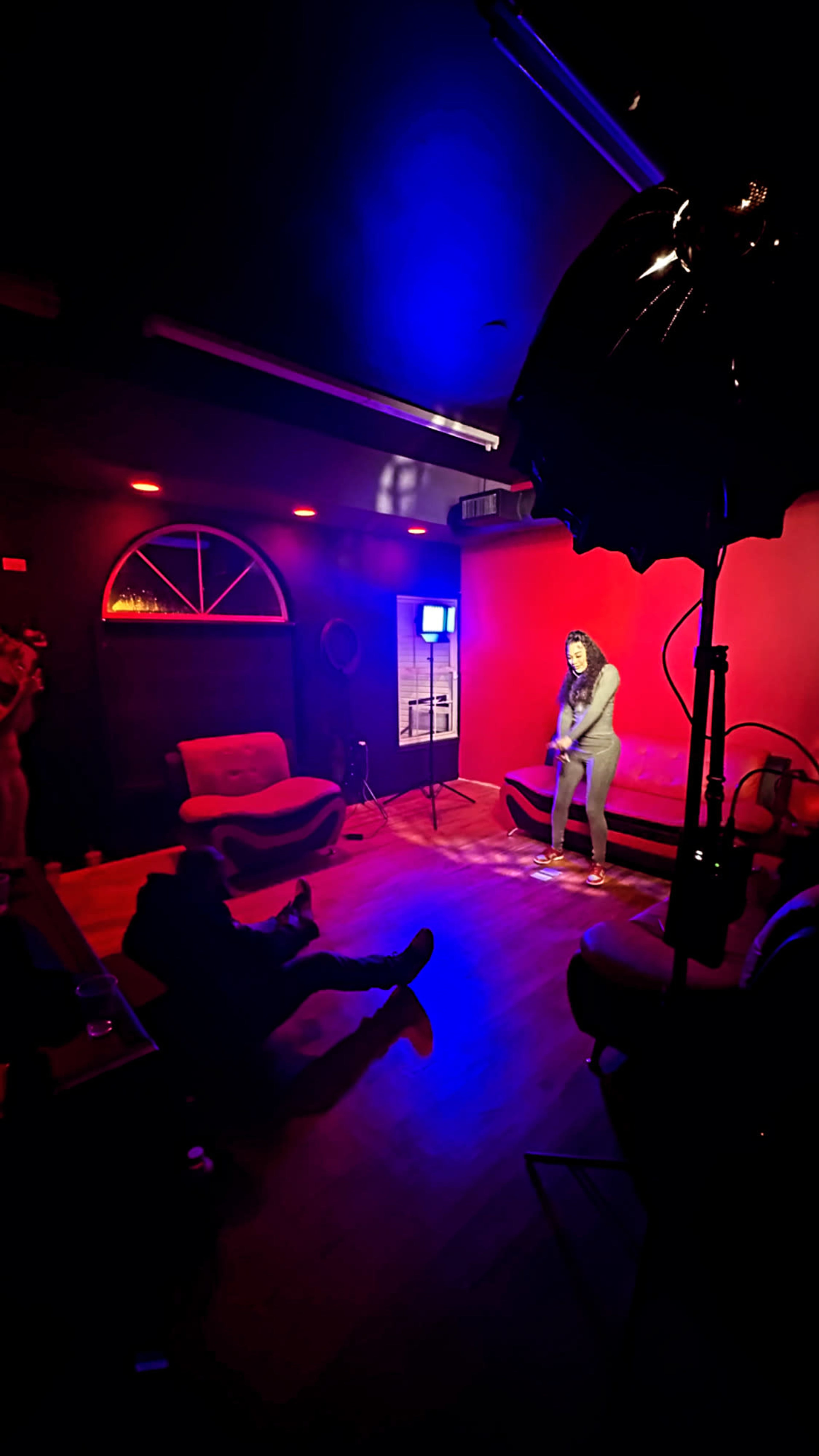 A performer stands on a small stage in a dimly lit room with red walls, while an audience sits on couches, observing the act.