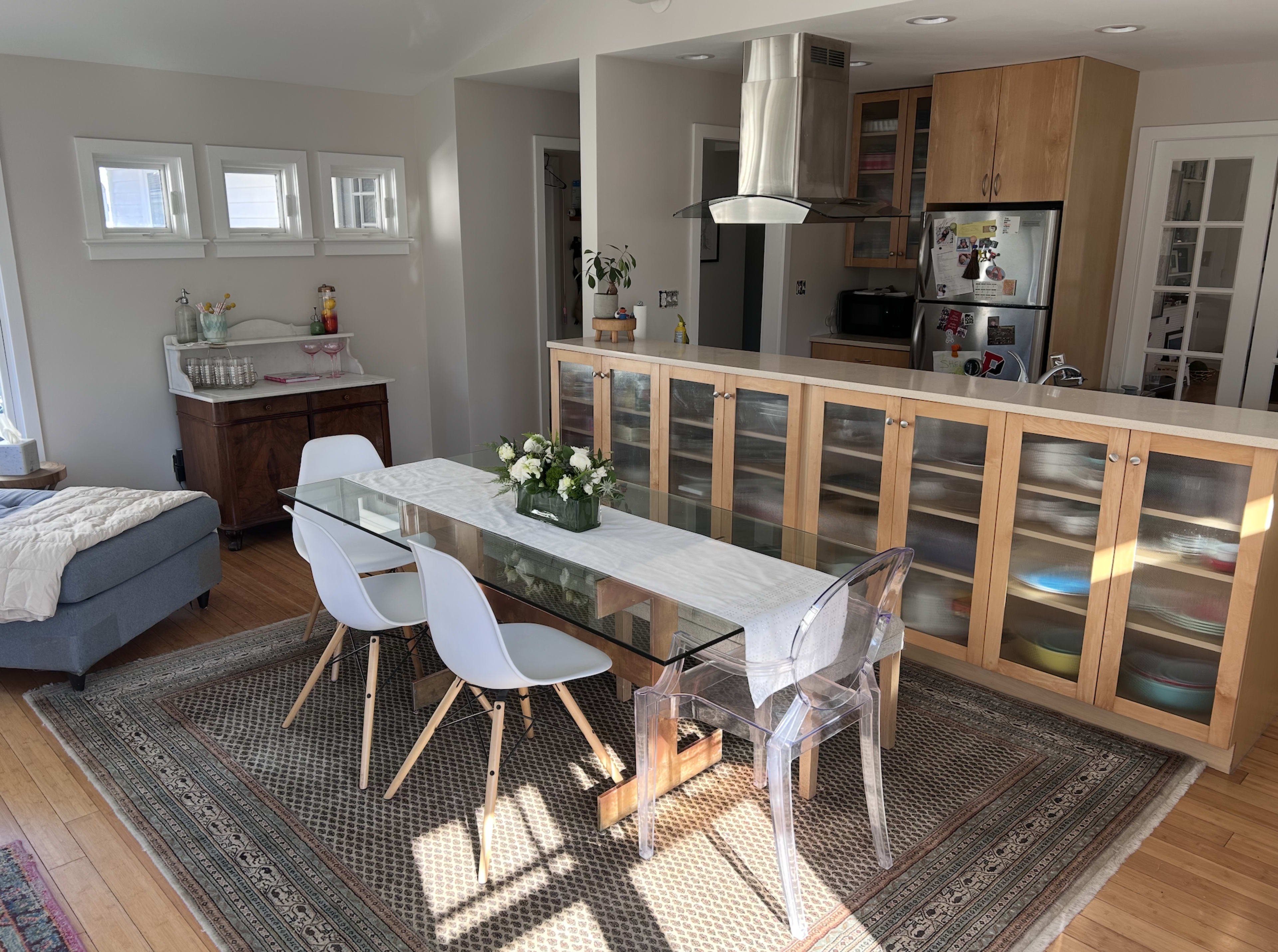 The image shows a modern dining area with a glass table and white chairs, connected to an open kitchen featuring wooden cabinets and stainless steel appliances.