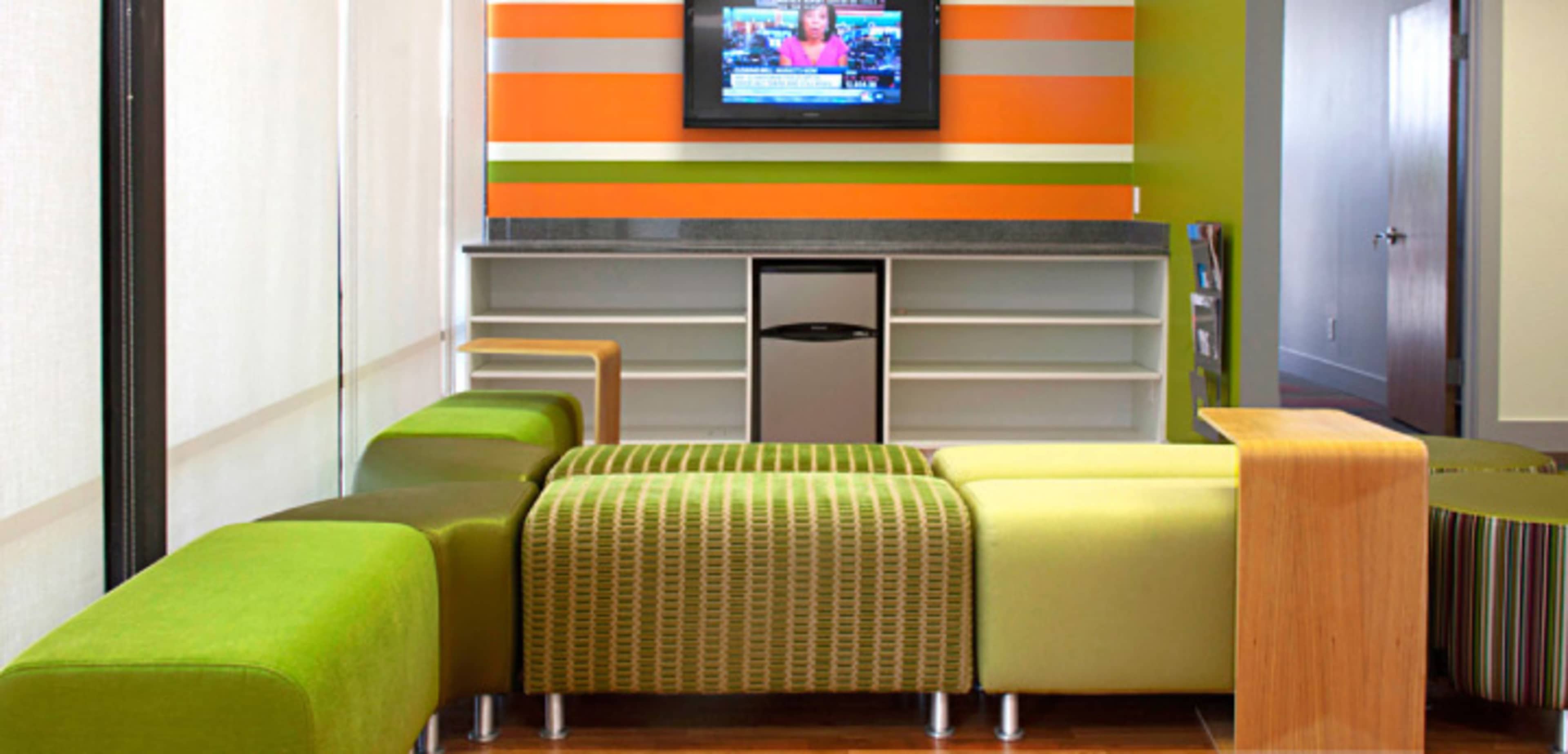 A modern seating area with green and striped couches, a television mounted on the wall, and a countertop with a small refrigerator in a brightly colored interior.
