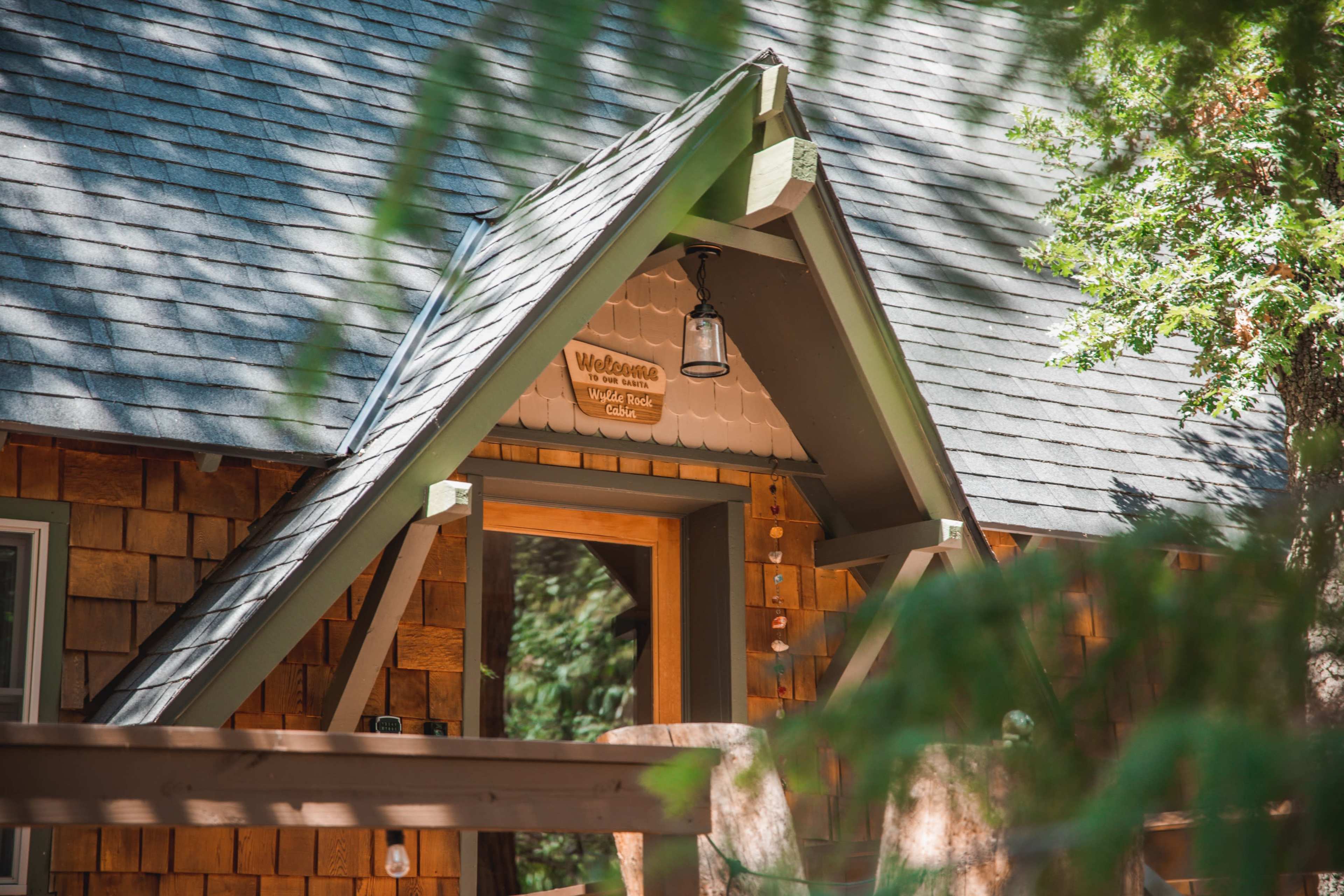 The image shows a cabin entrance with a triangular roof, wooden shingles, and a welcome sign above the door, surrounded by trees.