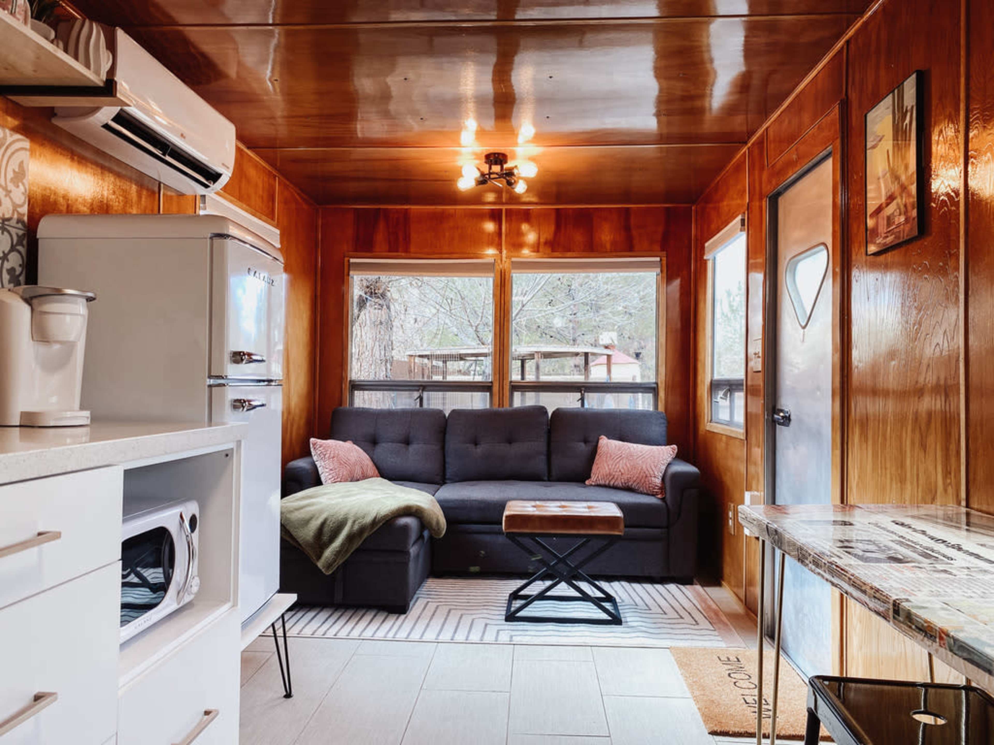 The image shows a cozy cabin interior featuring a living area with a dark sectional sofa, a wooden coffee table, and large windows that provide natural light.