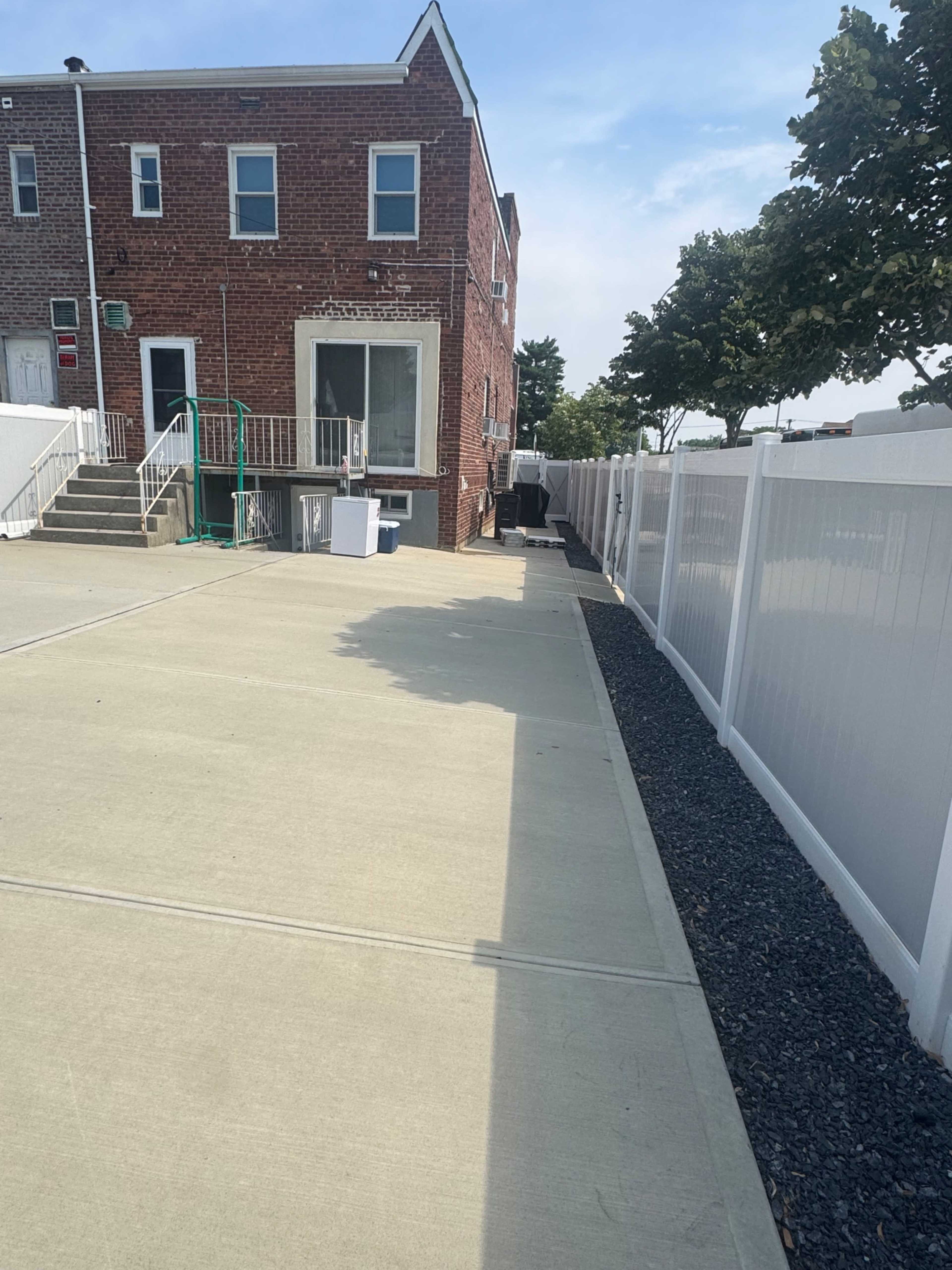 The image shows a concrete pathway beside a brick building, with a fenced area and several trees in the background.
