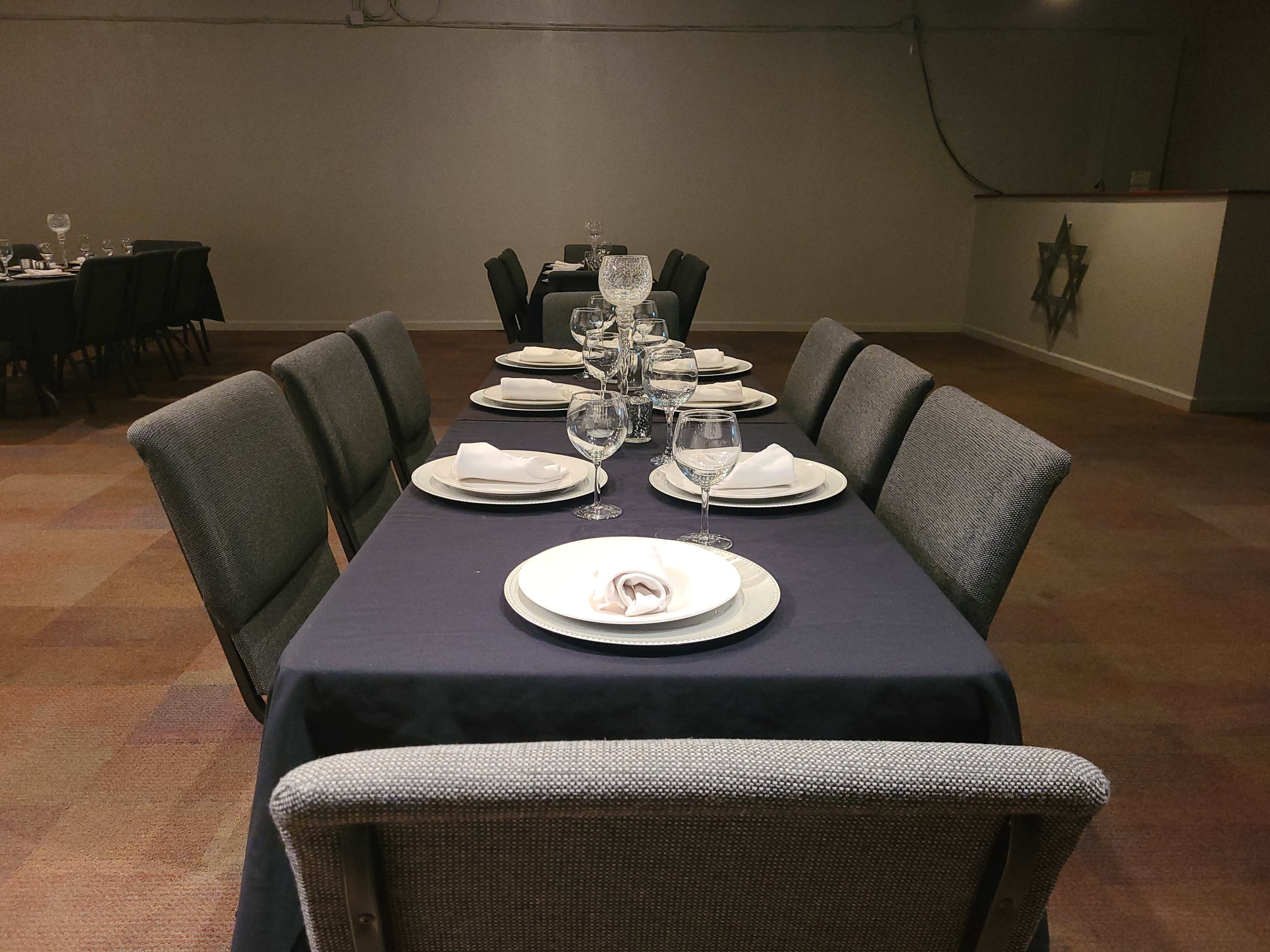 A long dining table is set with white plates and glassware, surrounded by gray upholstered chairs in a room with minimal decor.