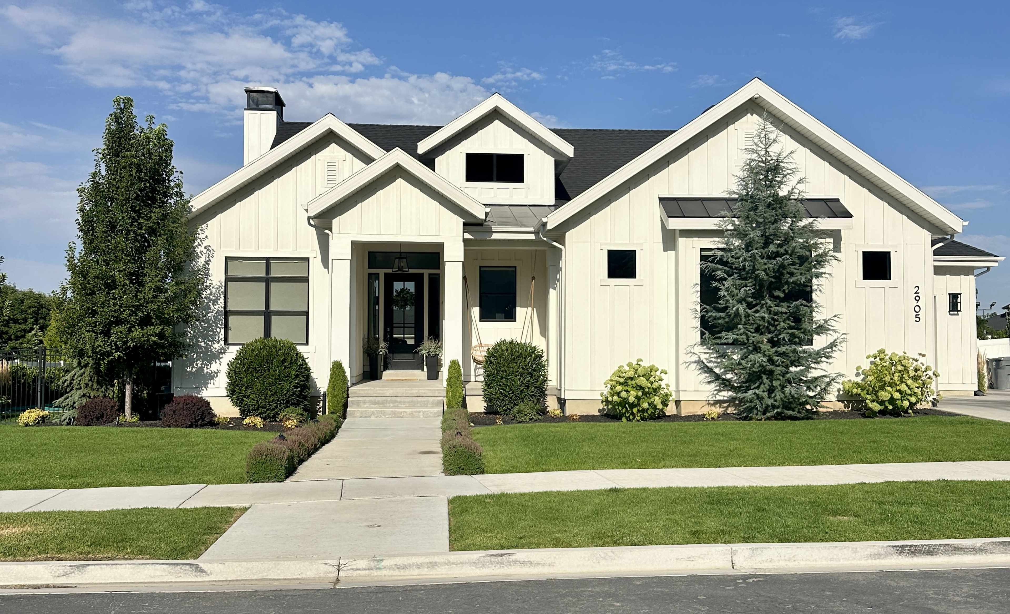 The image shows a modern, white-clapboard house with a landscaped front yard, a walkway leading to the entrance, and a blue sky overhead.