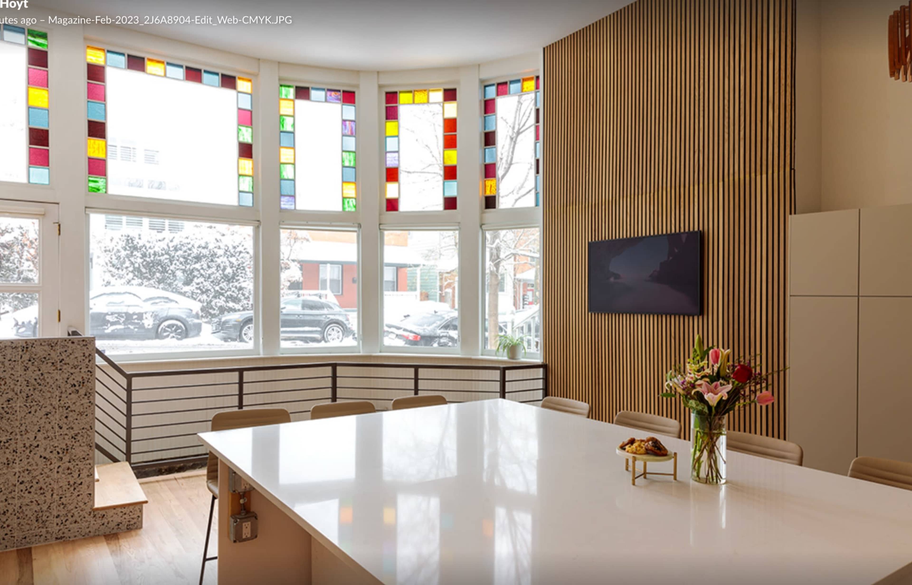 A spacious room features a large white table surrounded by chairs, with colorful stained glass windows and a view of a snowy landscape outside.