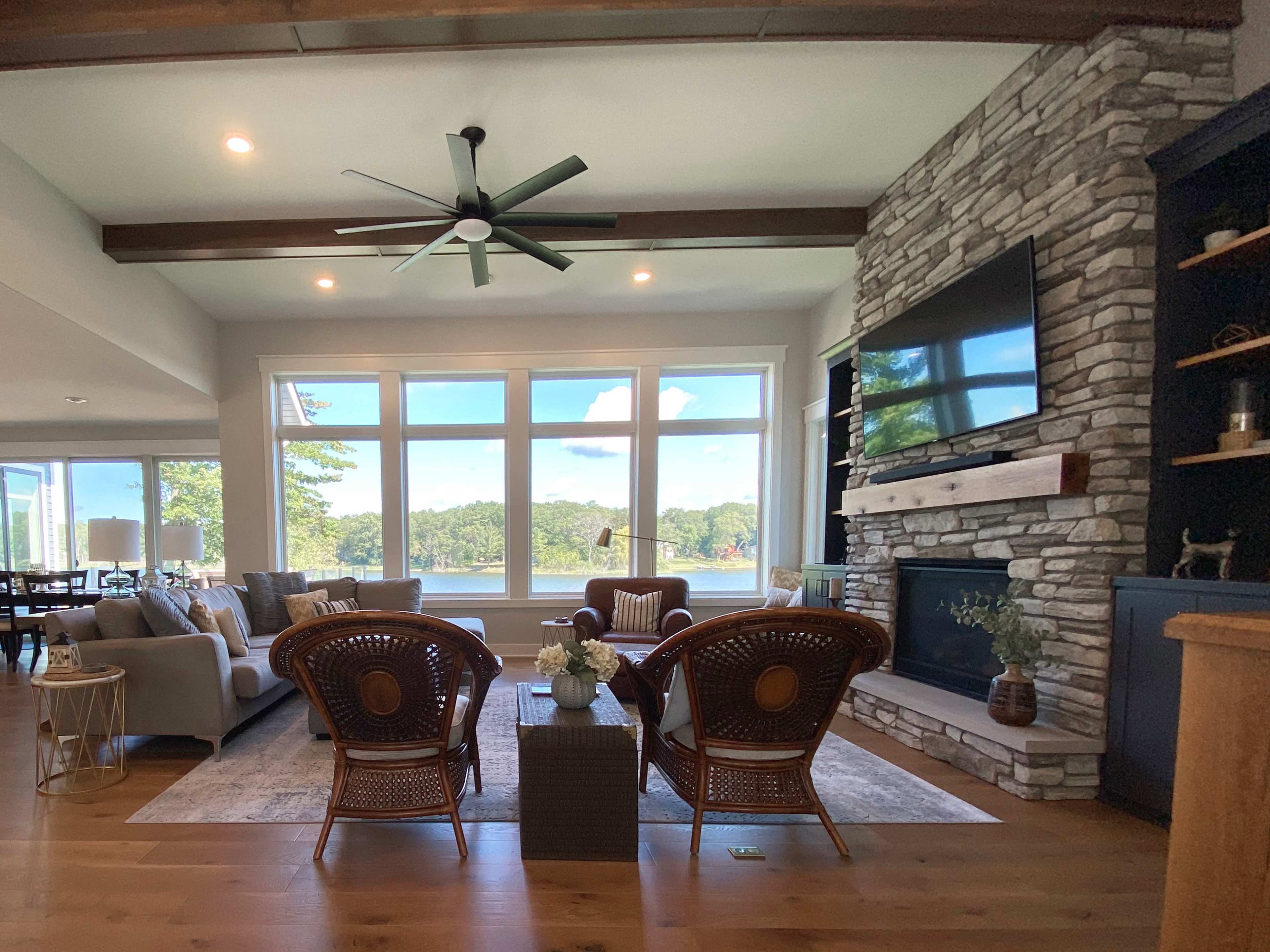 The living room features a stone fireplace and a seating area with wicker chairs, offering a view of a lake through large windows.