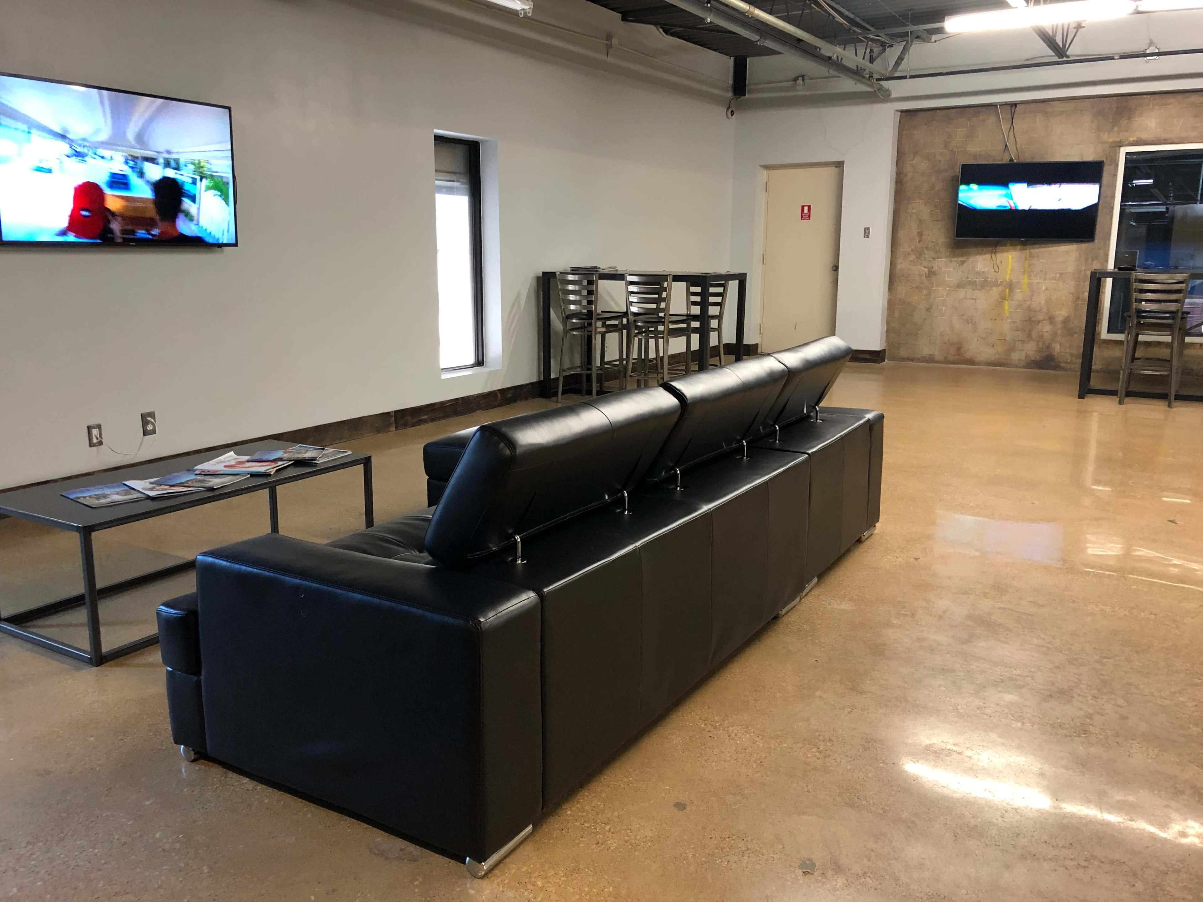 A black leather couch is positioned in a room with two televisions mounted on the walls and a table with chairs in the background.