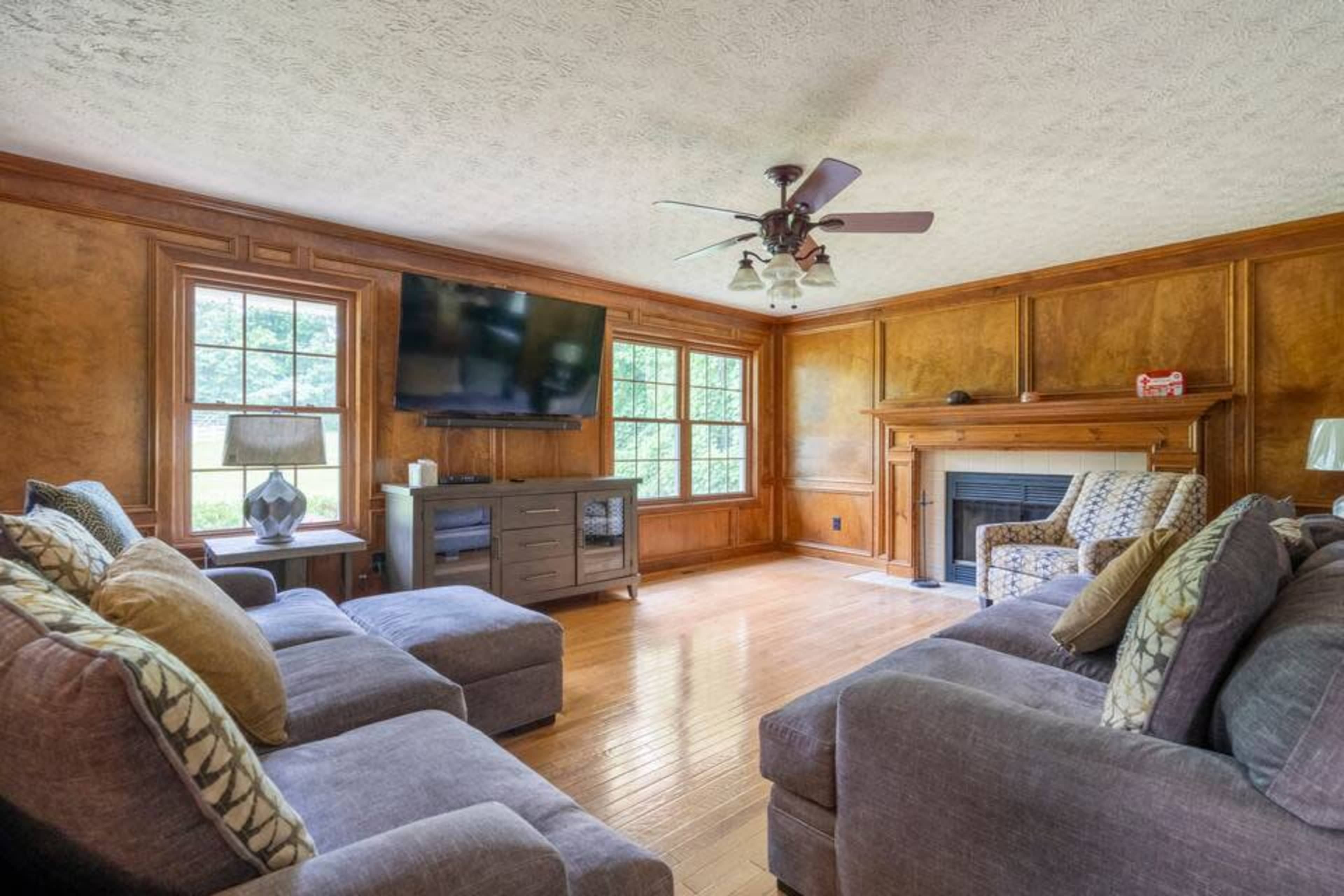 A living room features two sofas, a patterned armchair, a television mounted on the wall, and a wooden fireplace against a paneled backdrop.