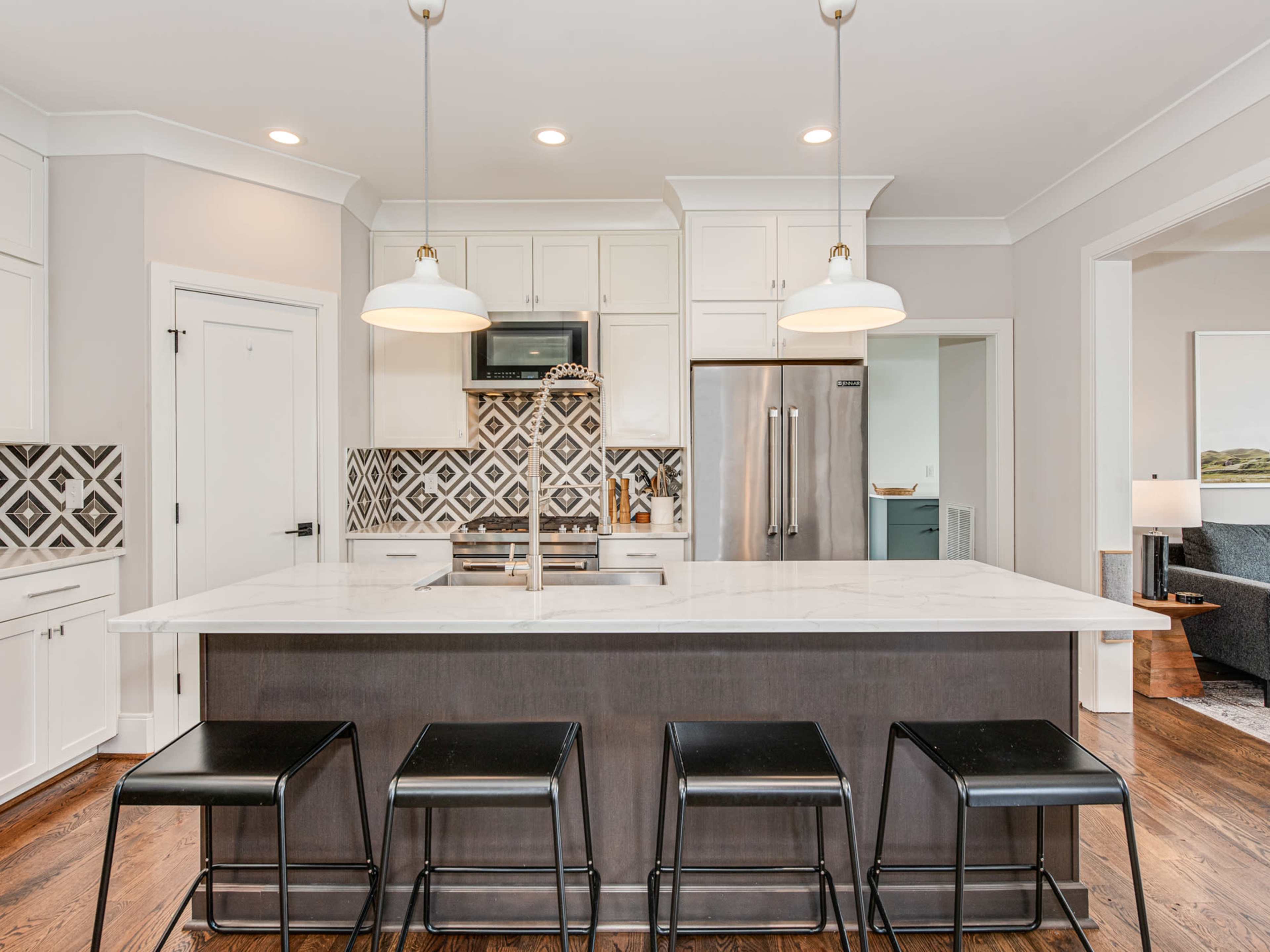 A modern kitchen features a large island with black stools, white cabinetry, and a patterned backsplash.