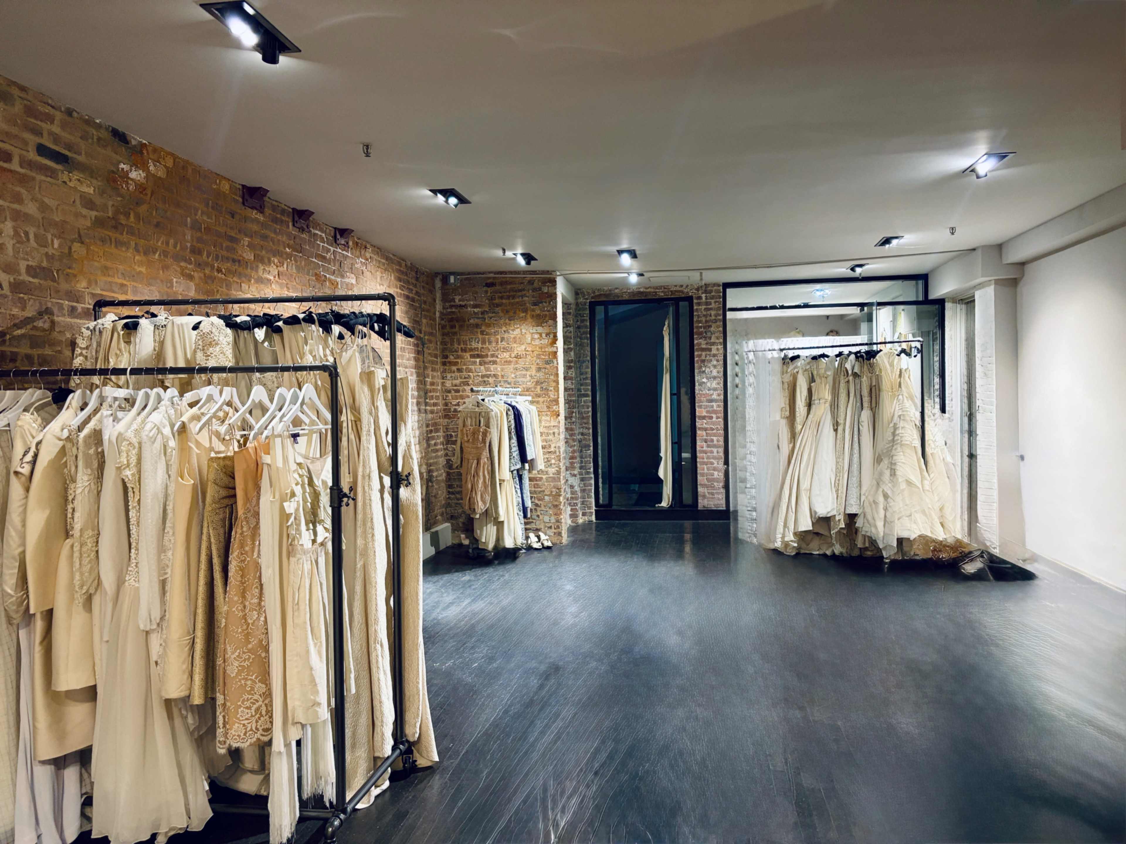 The image shows a spacious clothing store featuring racks of beige and cream-colored dresses against a brick wall.