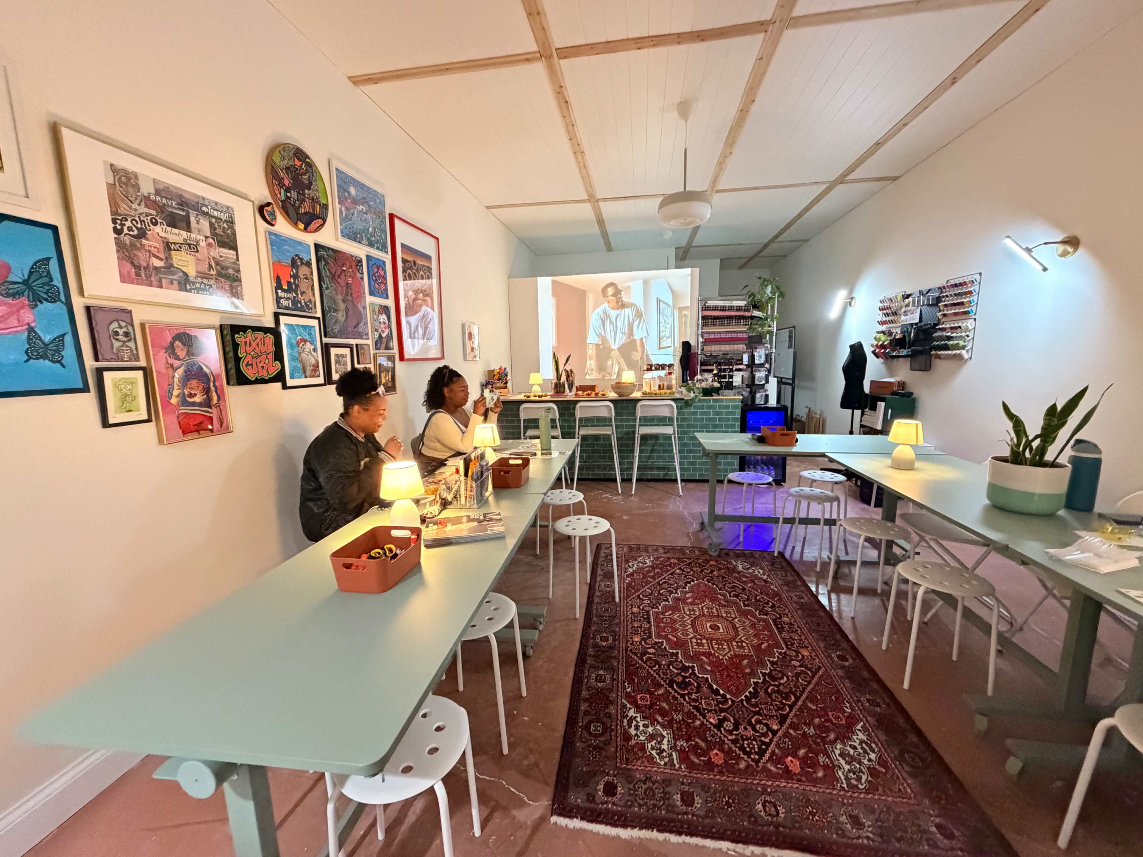 The image shows a spacious room with several tables and chairs, where two people are seated, and a projection featuring a person is visible on a wall.
