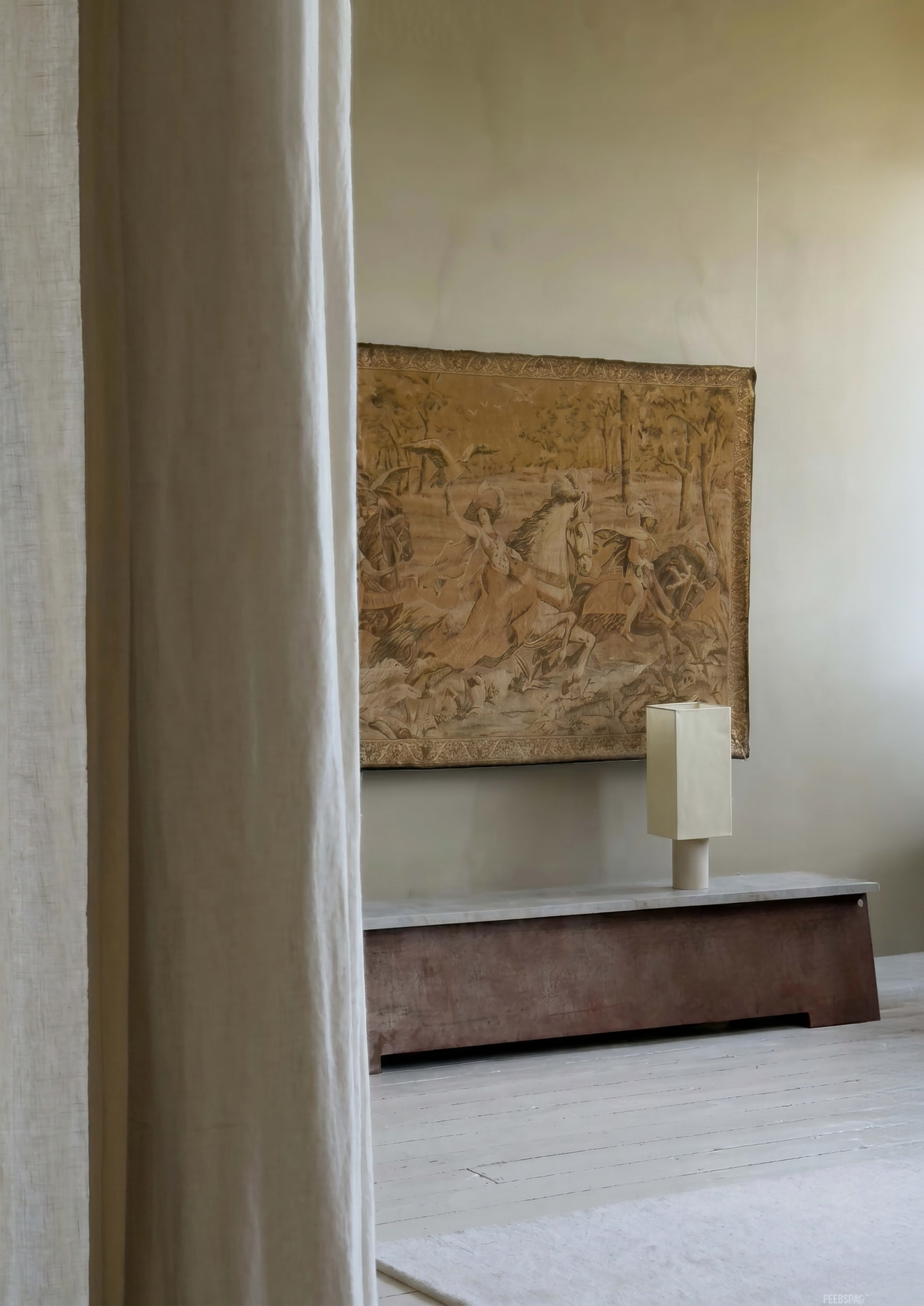 A textured tapestry depicting a pastoral scene hangs on a beige wall above a wooden bench with a minimalist lamp on top.