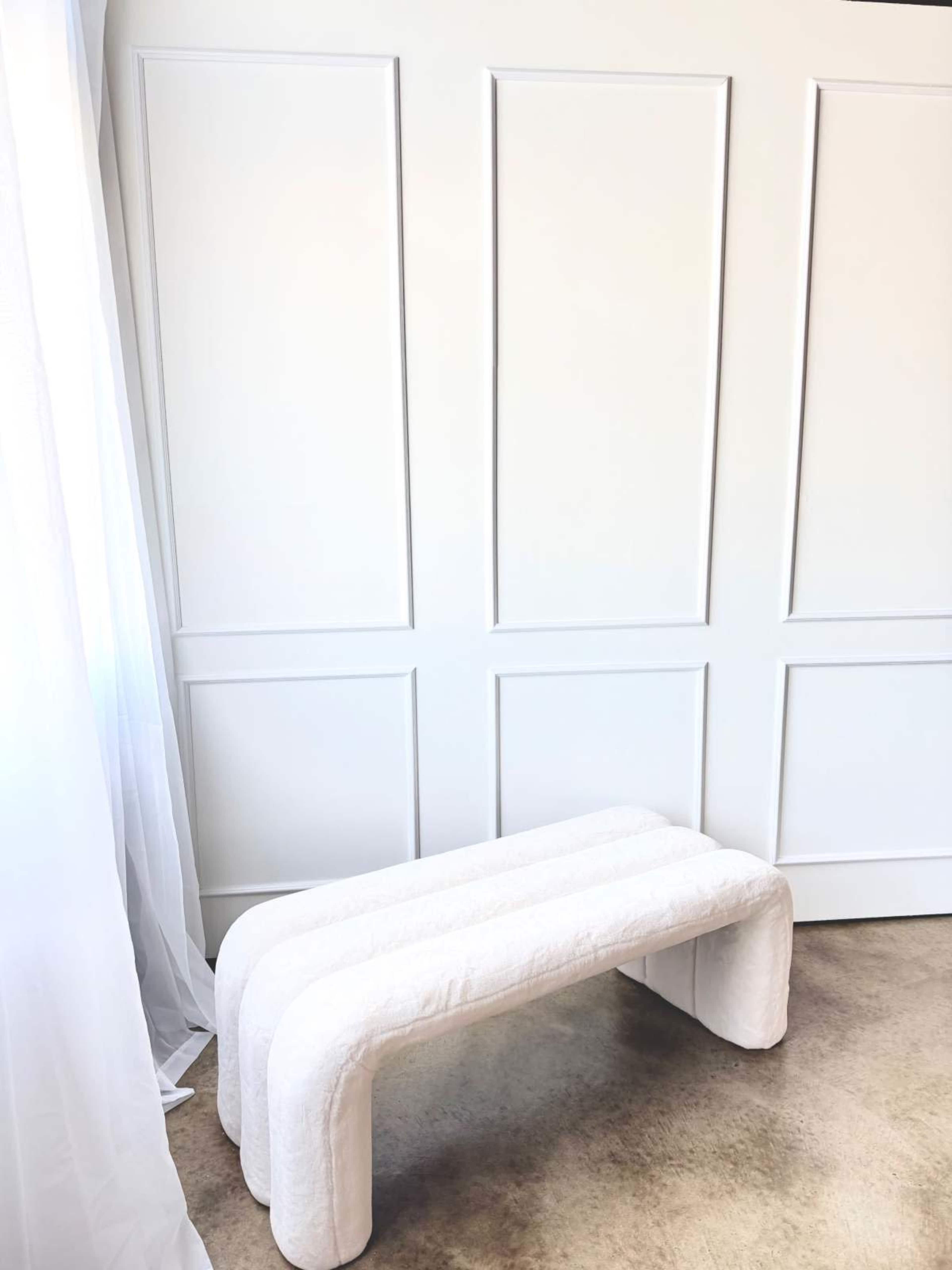 A white, upholstered bench with a rounded design is positioned against a paneled white wall.