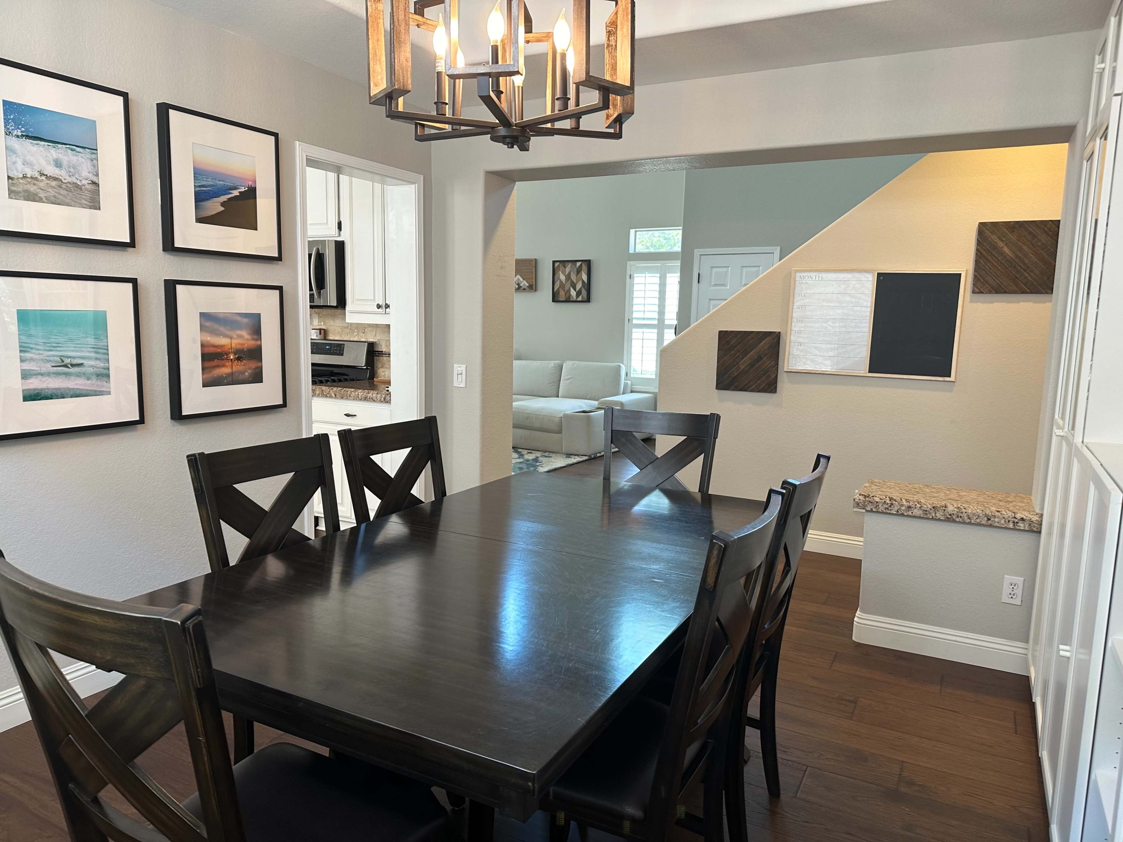 A dining area with a dark wooden table and chairs, framed photographs on the walls, and a view into a living room through an open archway.
