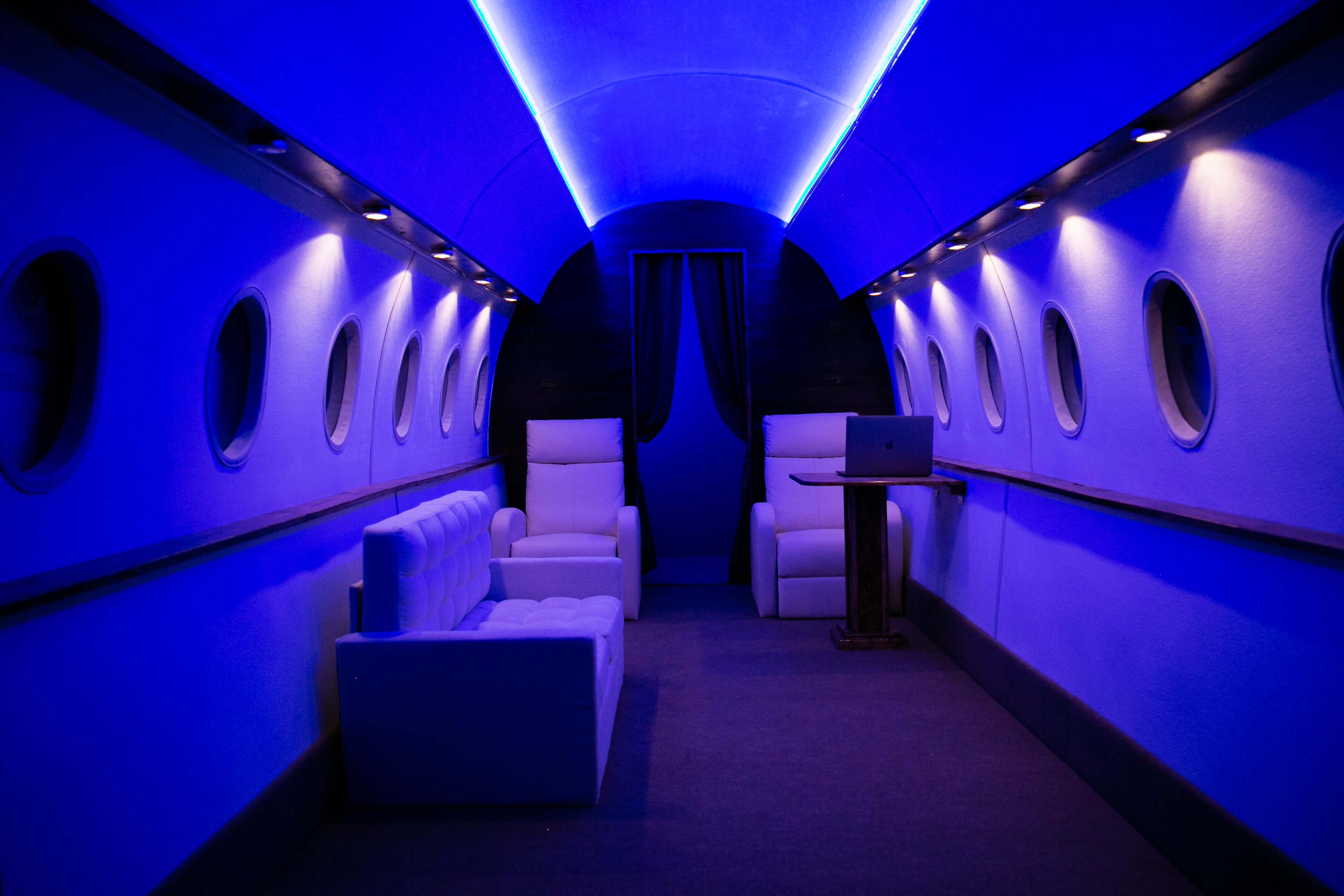 The interior of a jet is illuminated with blue lighting and features white seating arrangements, including a sofa and armchairs.