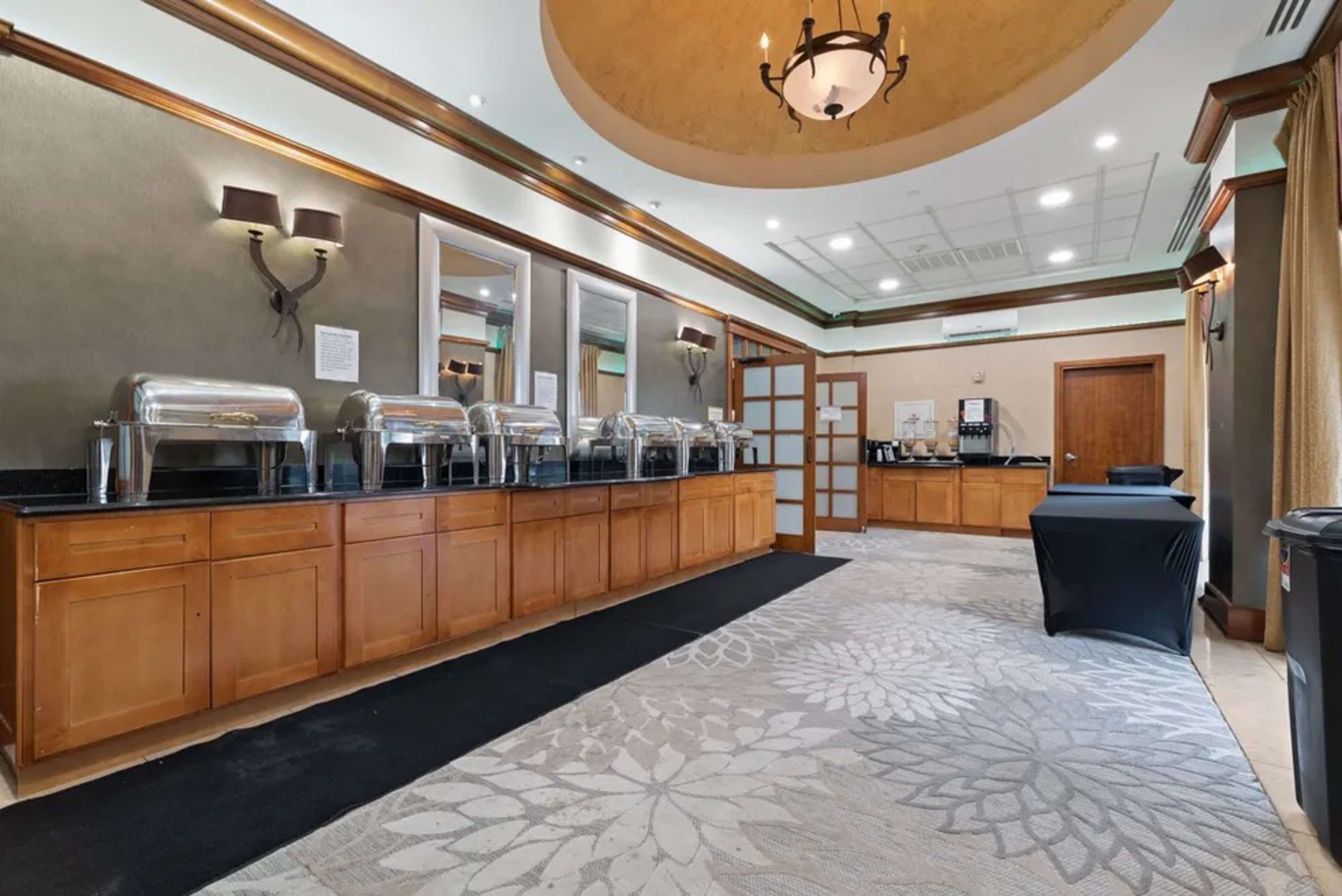 A buffet area with several food warmers along a wooden counter, adjacent to a coffee station and a separate table draped in black cloth.