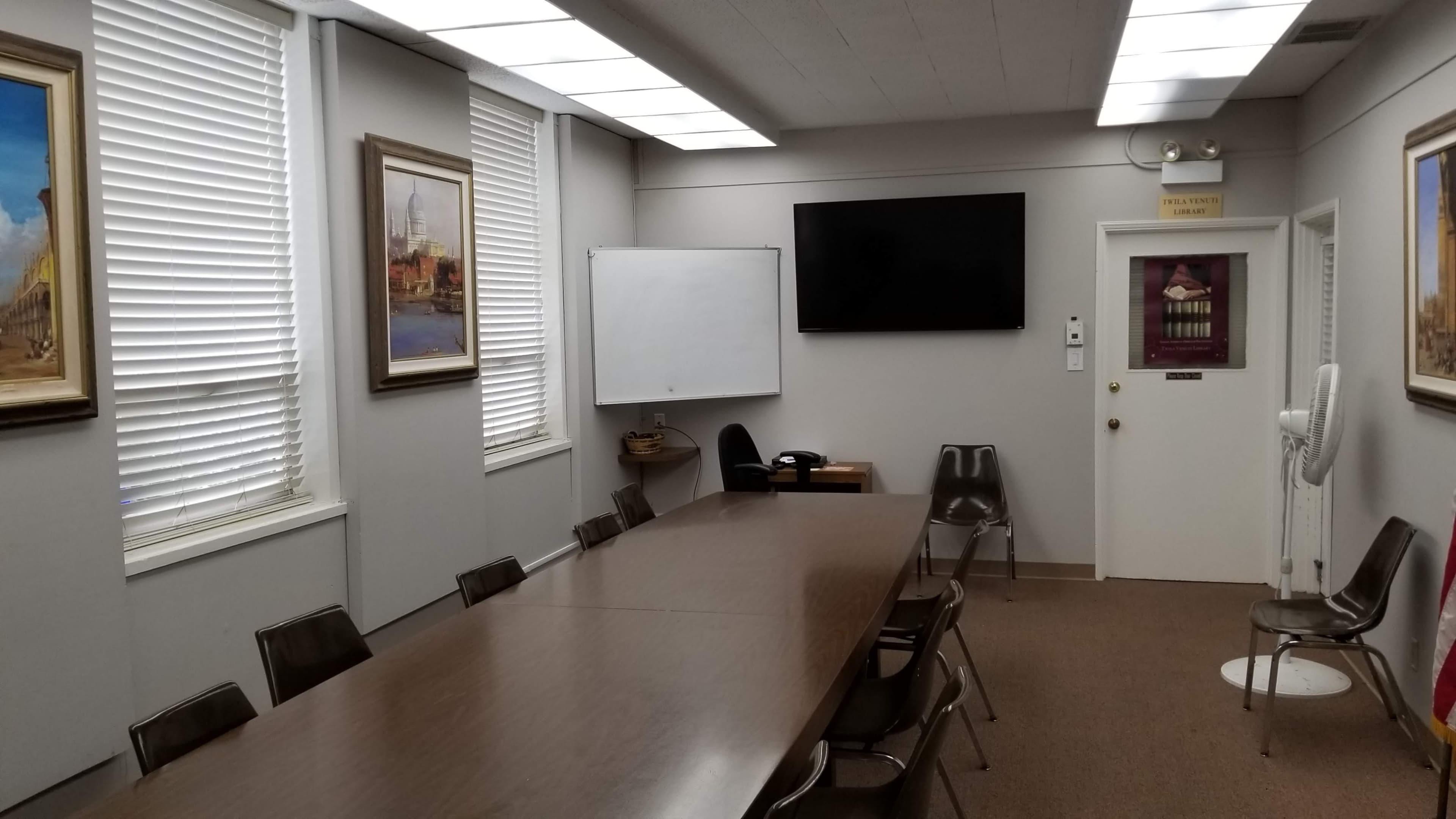 A conference room features a long wooden table, several chairs, a whiteboard, a television mounted on the wall, and framed paintings.