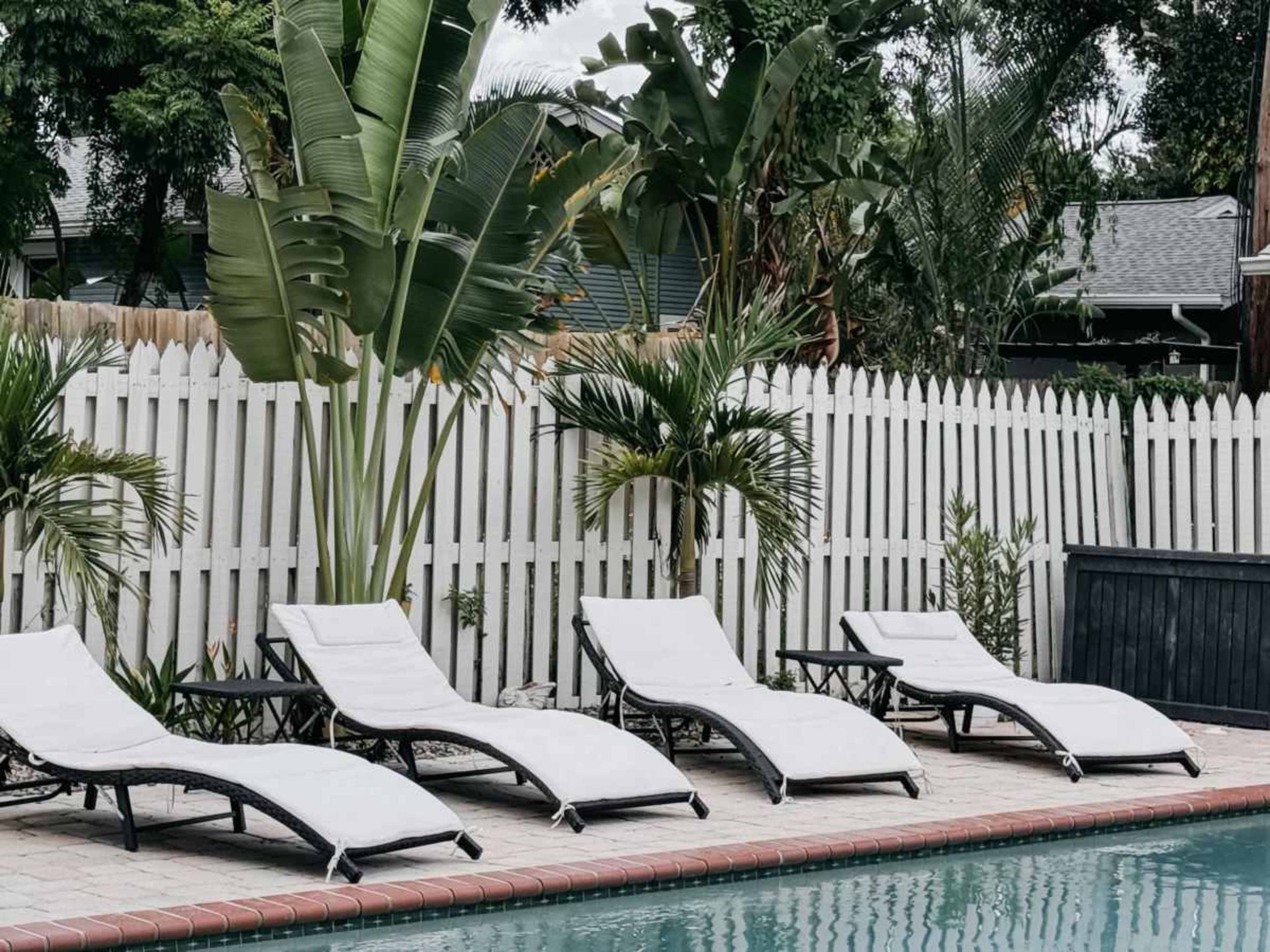 Four white lounge chairs are lined up beside a swimming pool, surrounded by greenery and a white picket fence.
