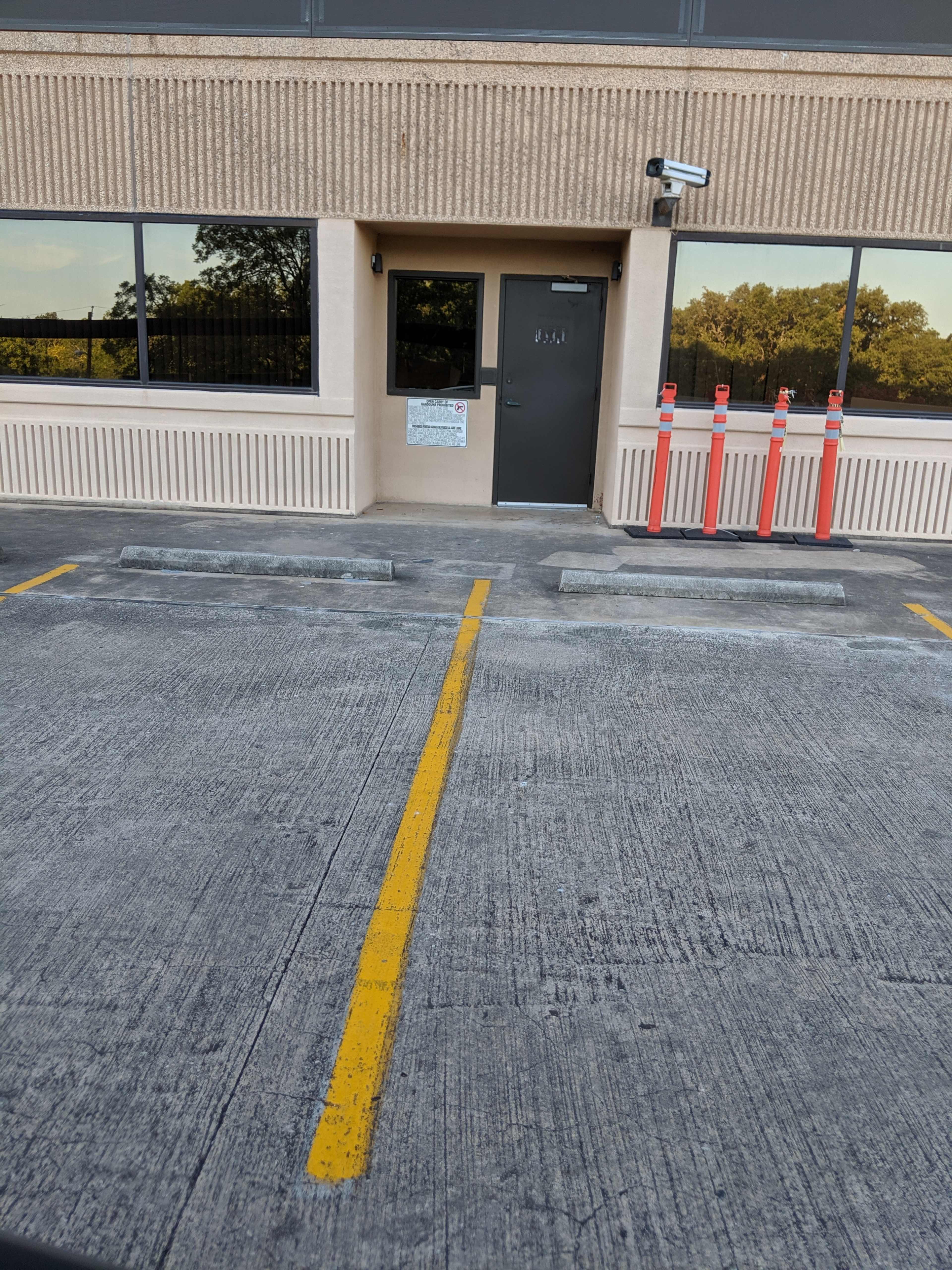 A building entrance with a closed door, surveillance camera, and empty parking spaces marked by a yellow line in front.