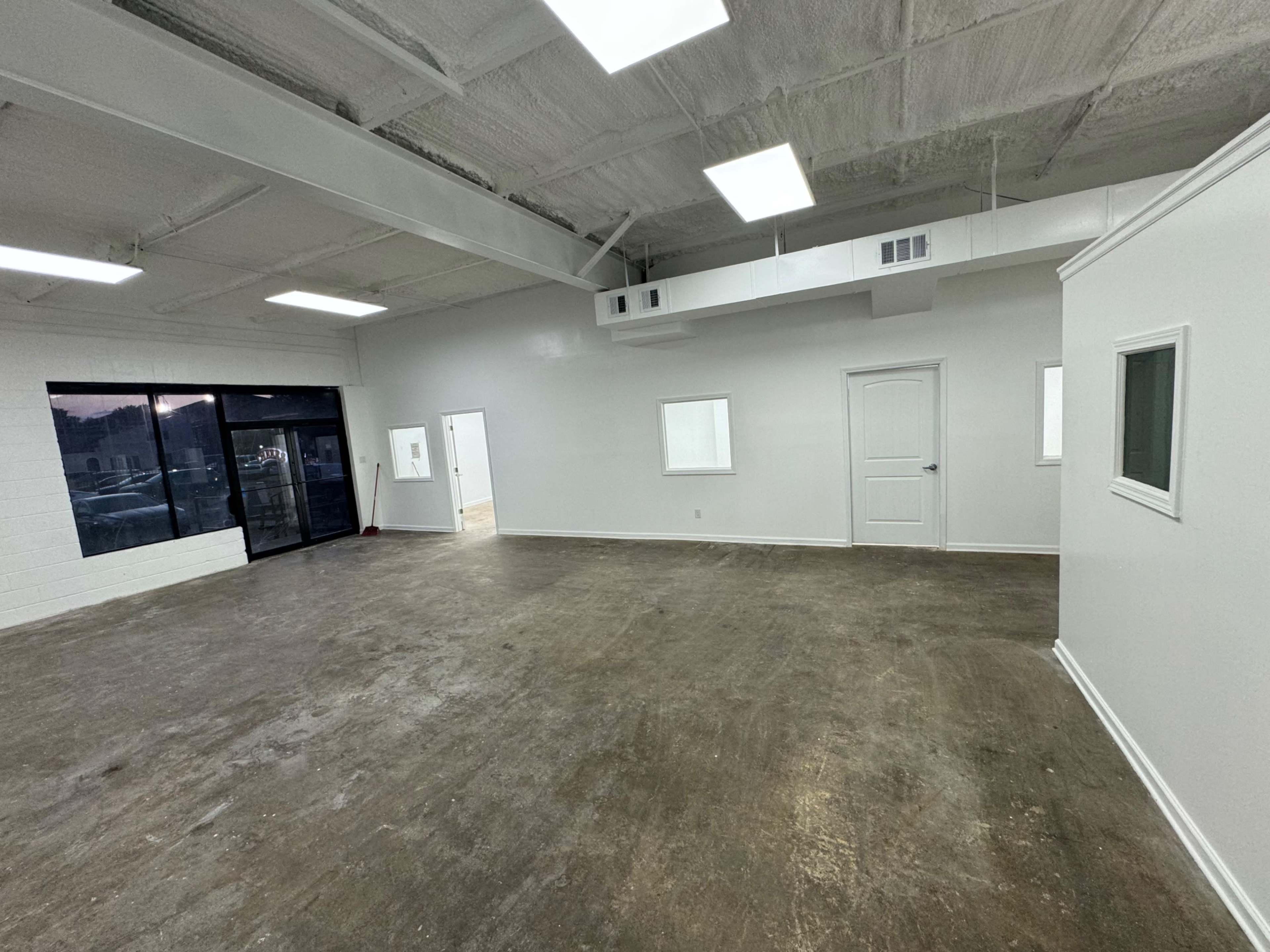 The image shows a spacious, empty commercial room with a concrete floor and white walls, featuring several windows and a single door.