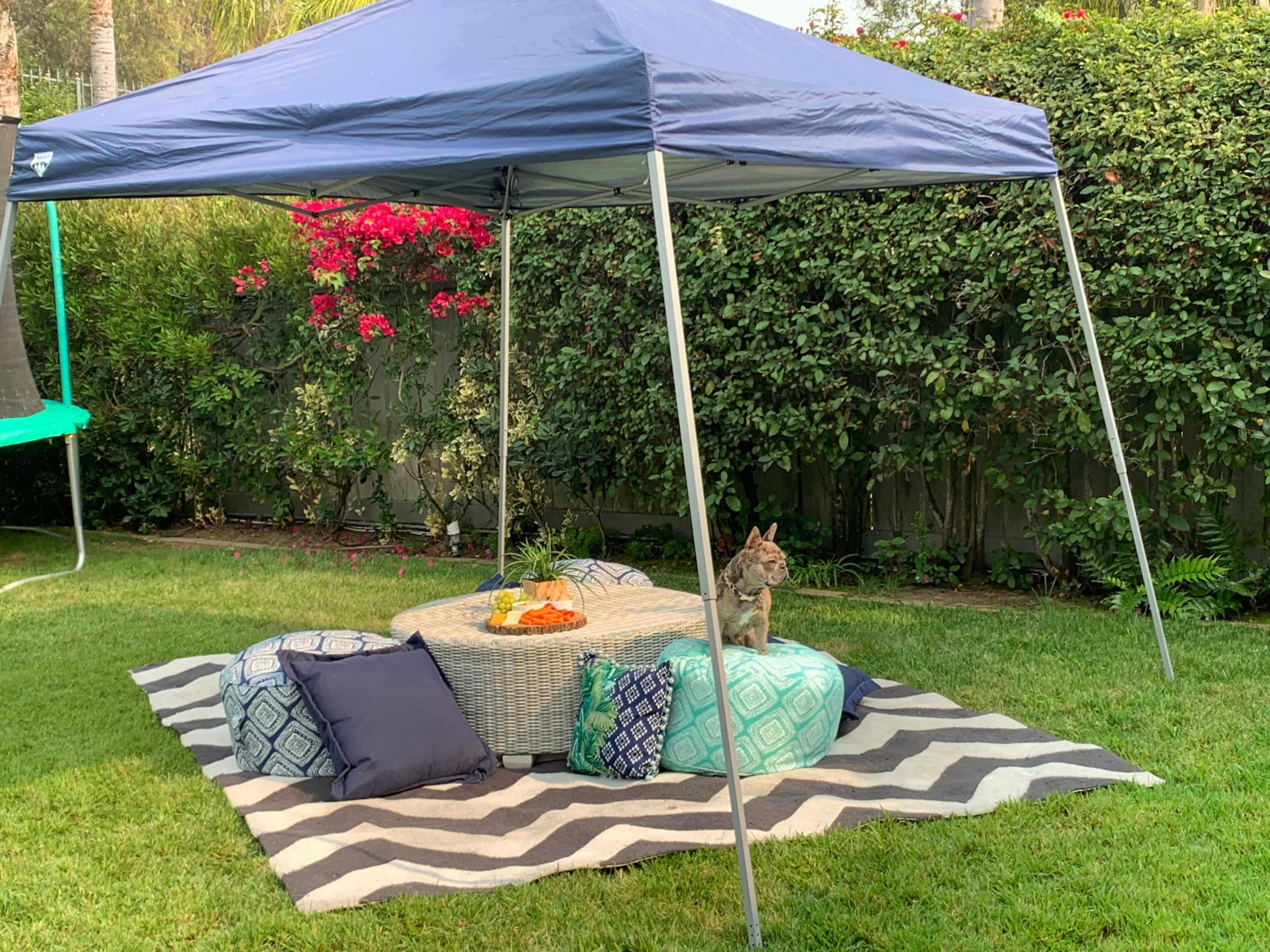 A tent covers a seating area with a table and colorful cushions on a grassy lawn surrounded by flowers and greenery.