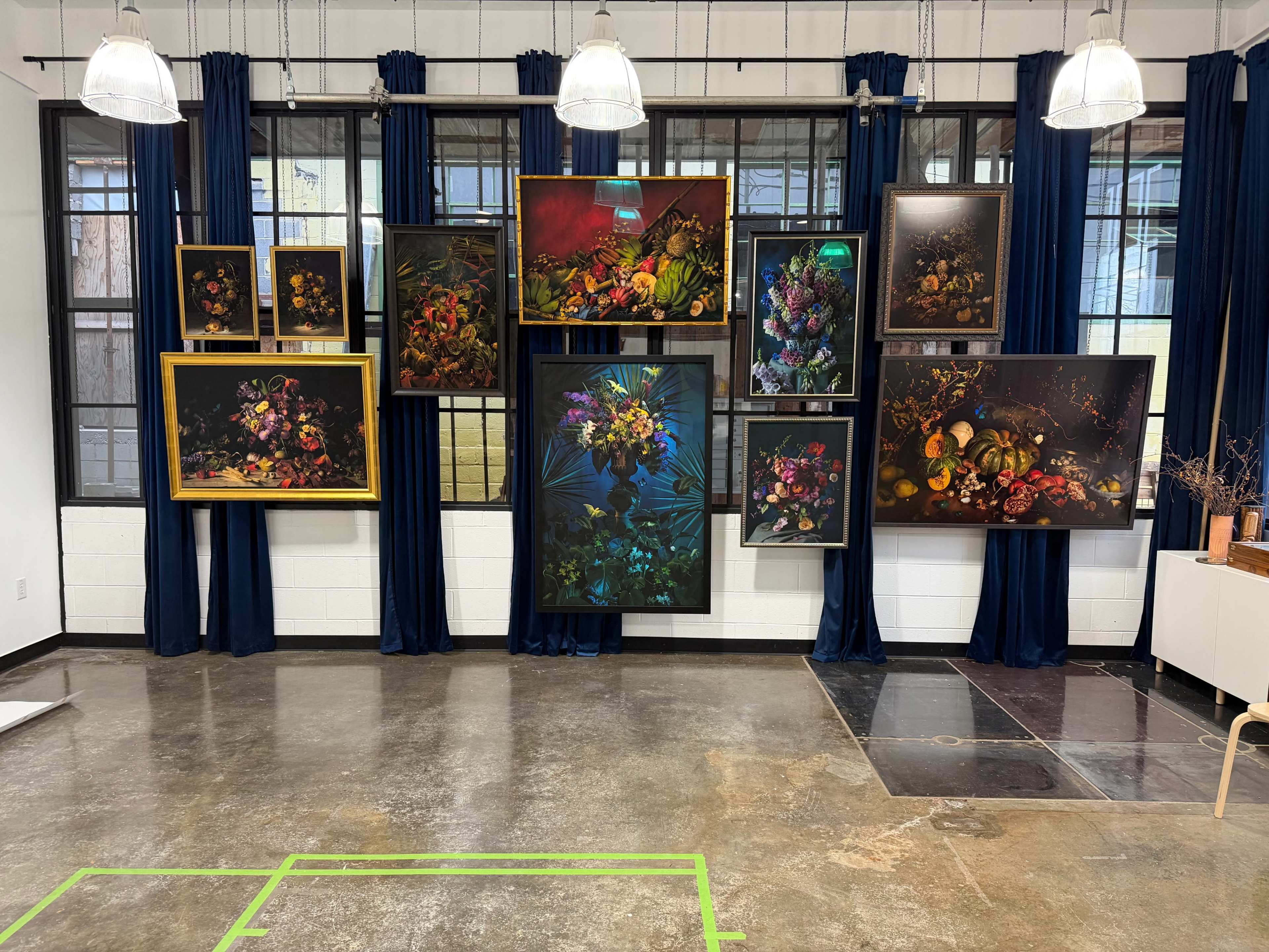 The image shows a gallery wall adorned with various framed floral artworks, set against dark blue curtains in a well-lit space.