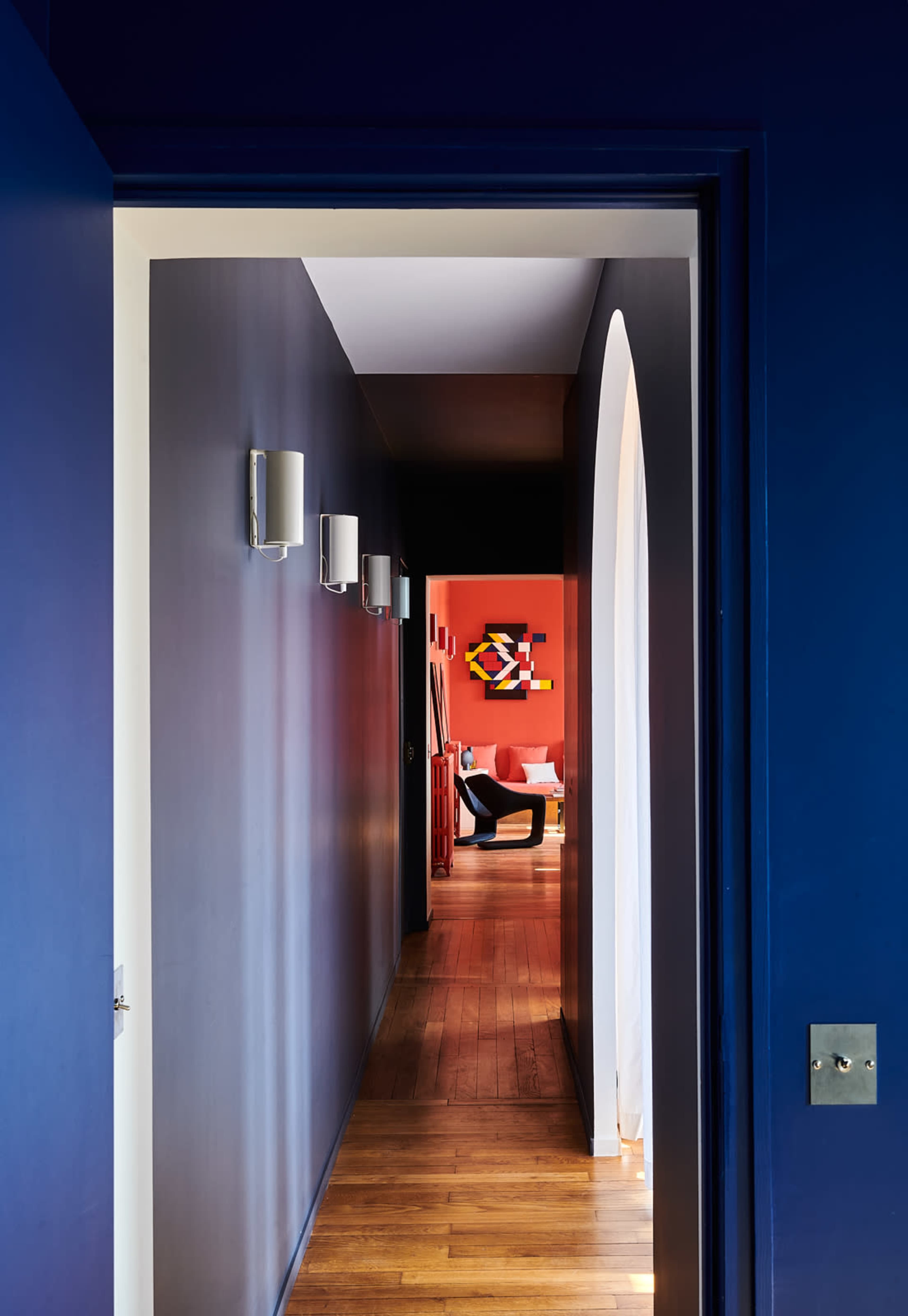 A hallway with dark blue walls leads to a brightly colored room featuring an abstract artwork on a red wall.