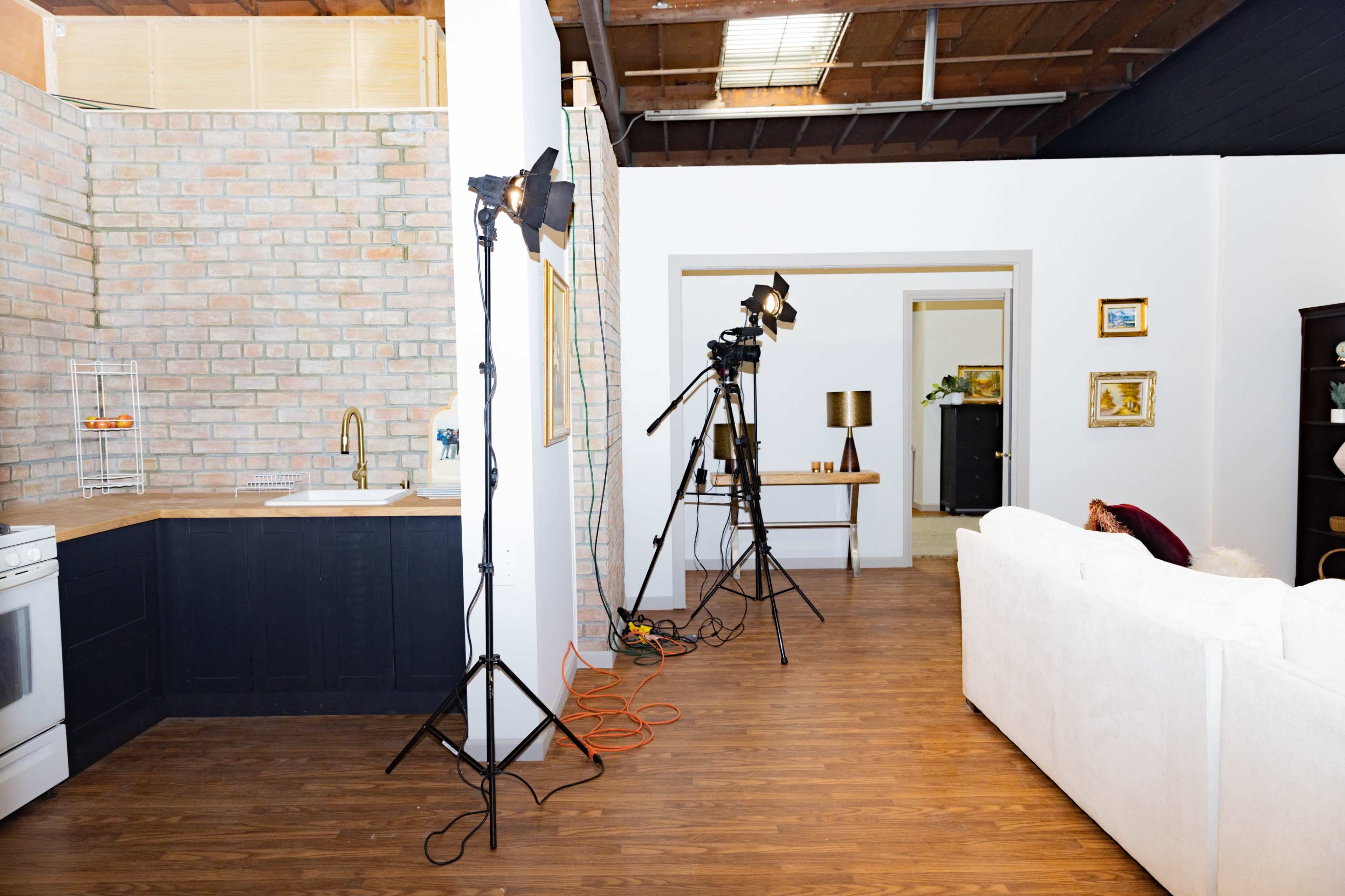 The scene features a studio set-up with a kitchenette, a white couch, and camera equipment positioned on tripods.