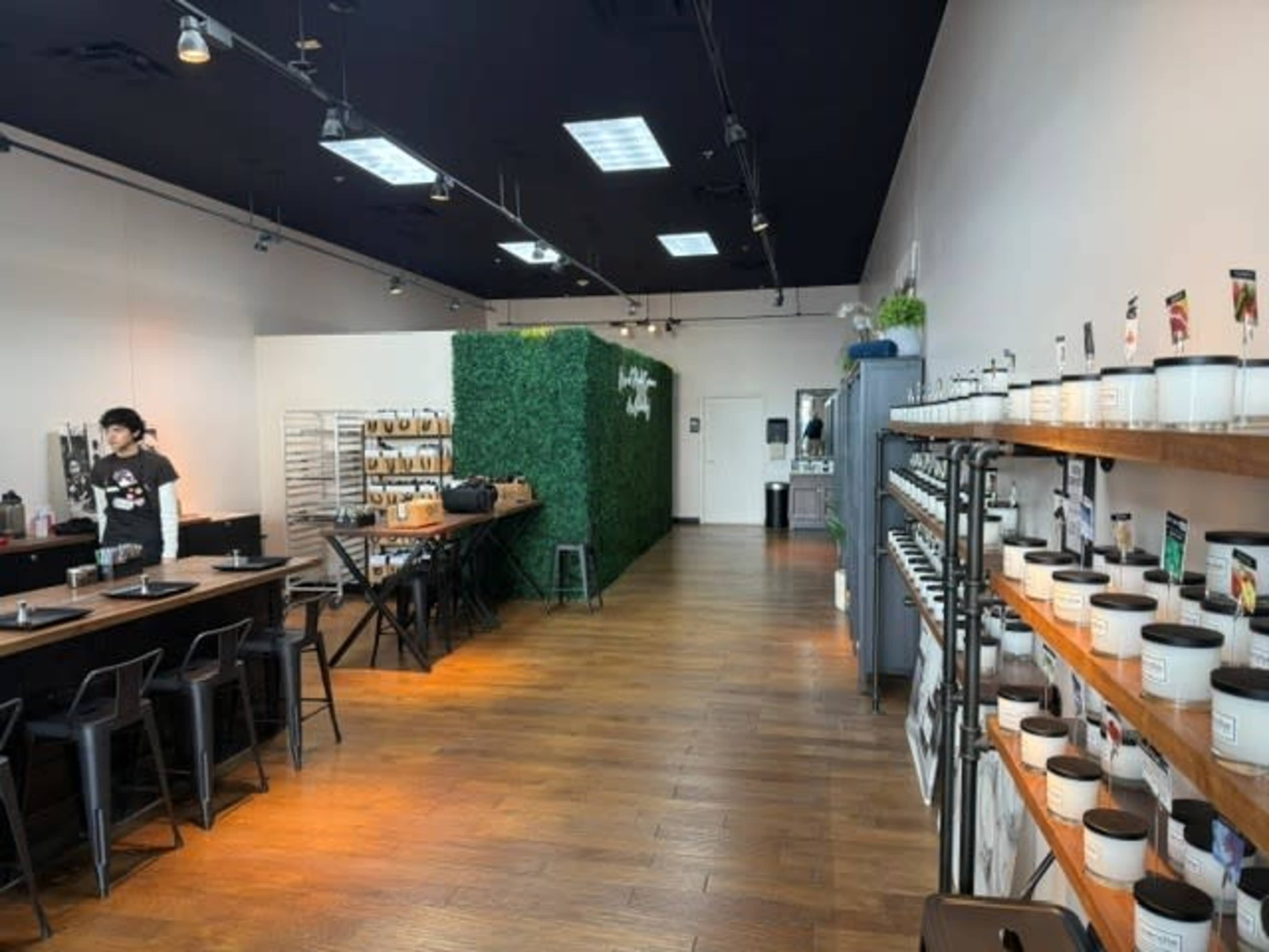 The image shows a modern cafe interior with a seating area on the left, a green plant wall in the center, and product displays along the right wall.